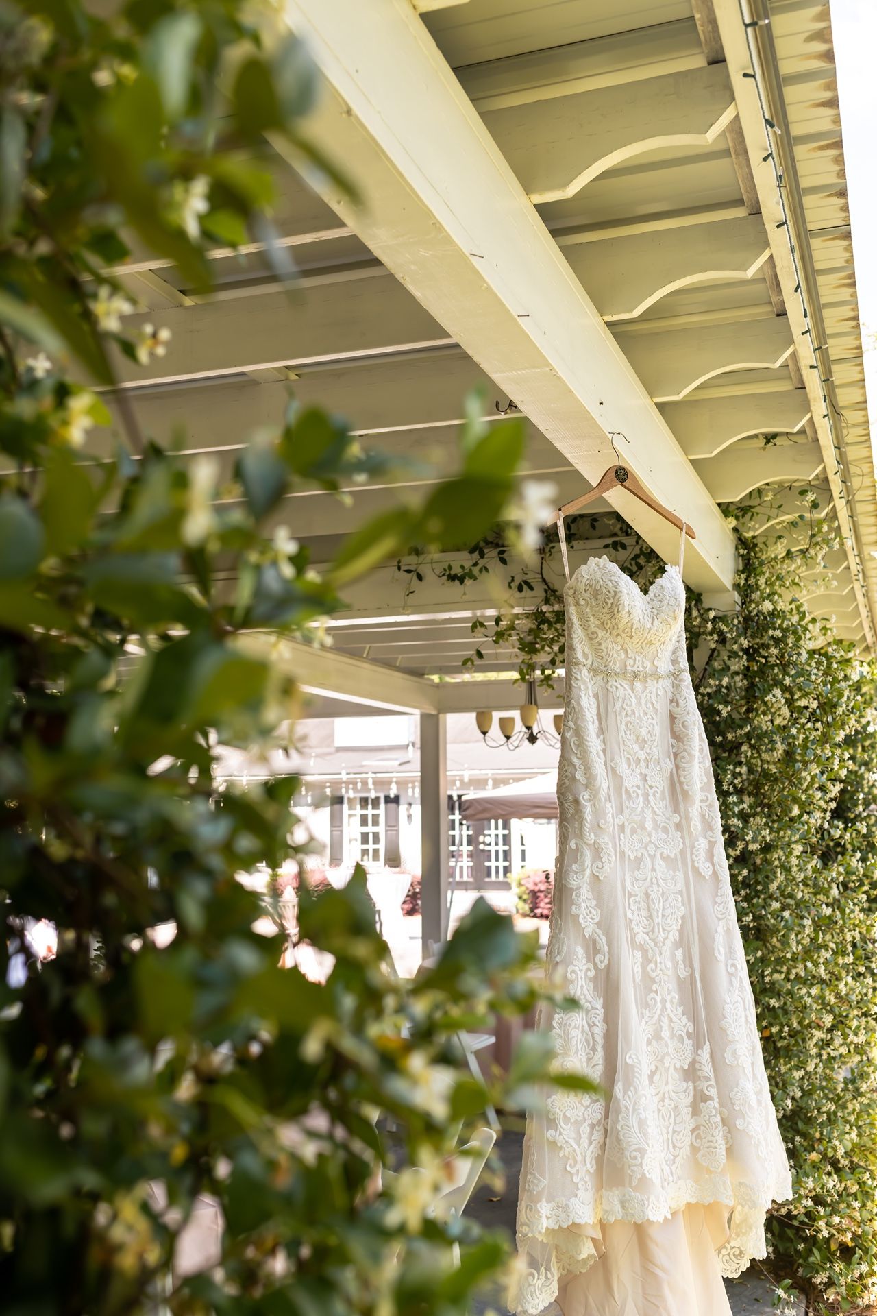 An elegant white lace wedding dress hangs from a porch rafter, framed by green foliage and soft natural lighting.