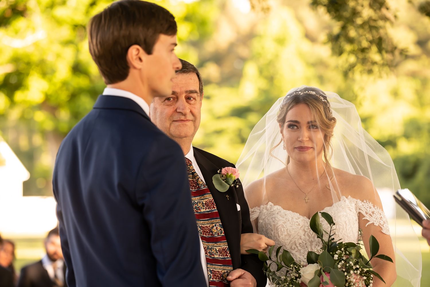 A wedding ceremony takes place outdoors with people standing together during the vow exchange on a sunny day.