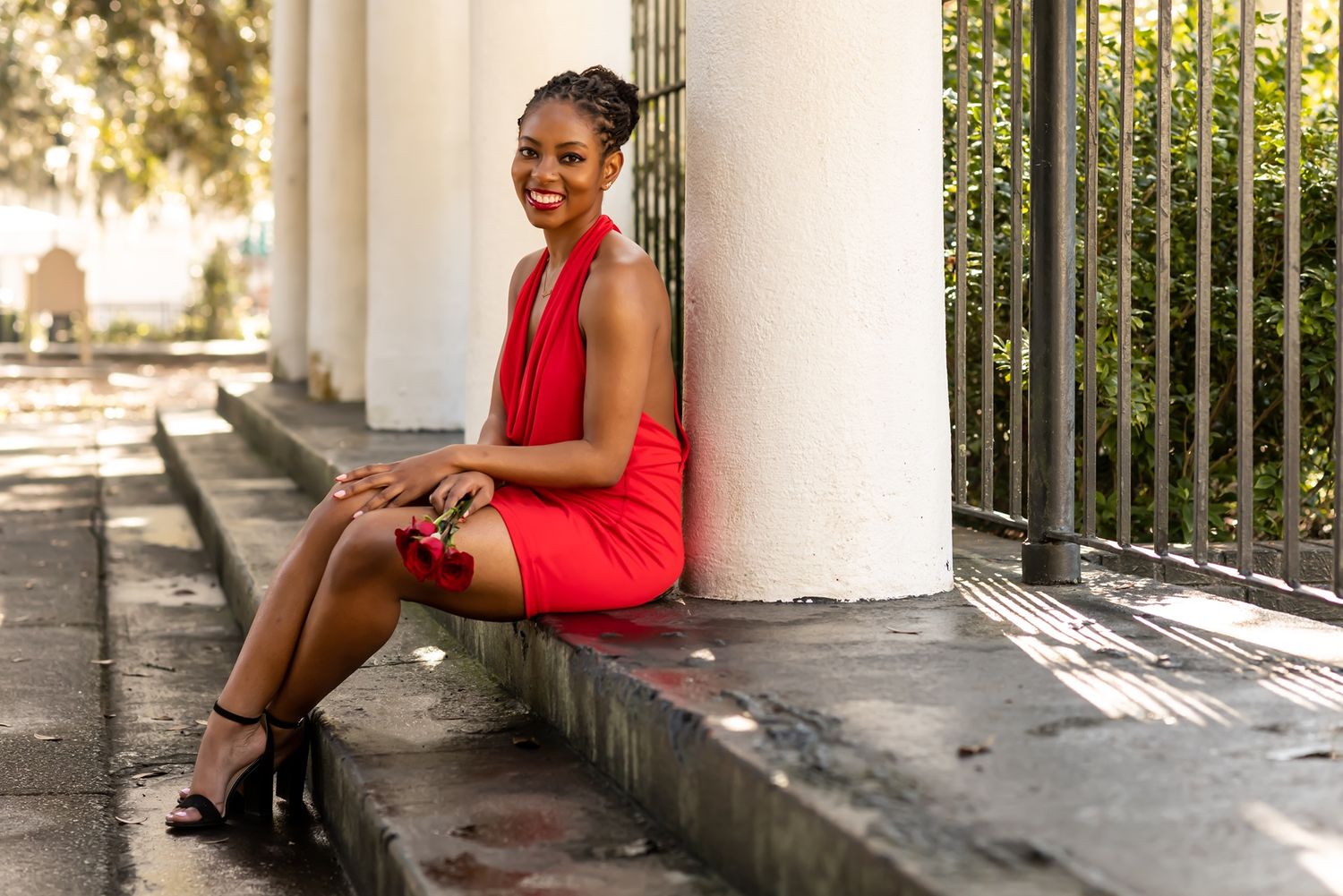 Person in a vibrant red dress sits elegantly on outdoor steps in a sunlit architectural setting.