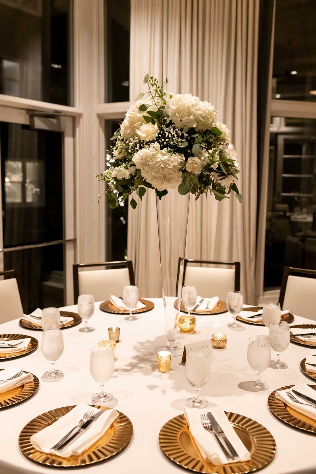 Elegant wedding reception table setting with tall white floral centerpiece, gold charger plates and white linens.