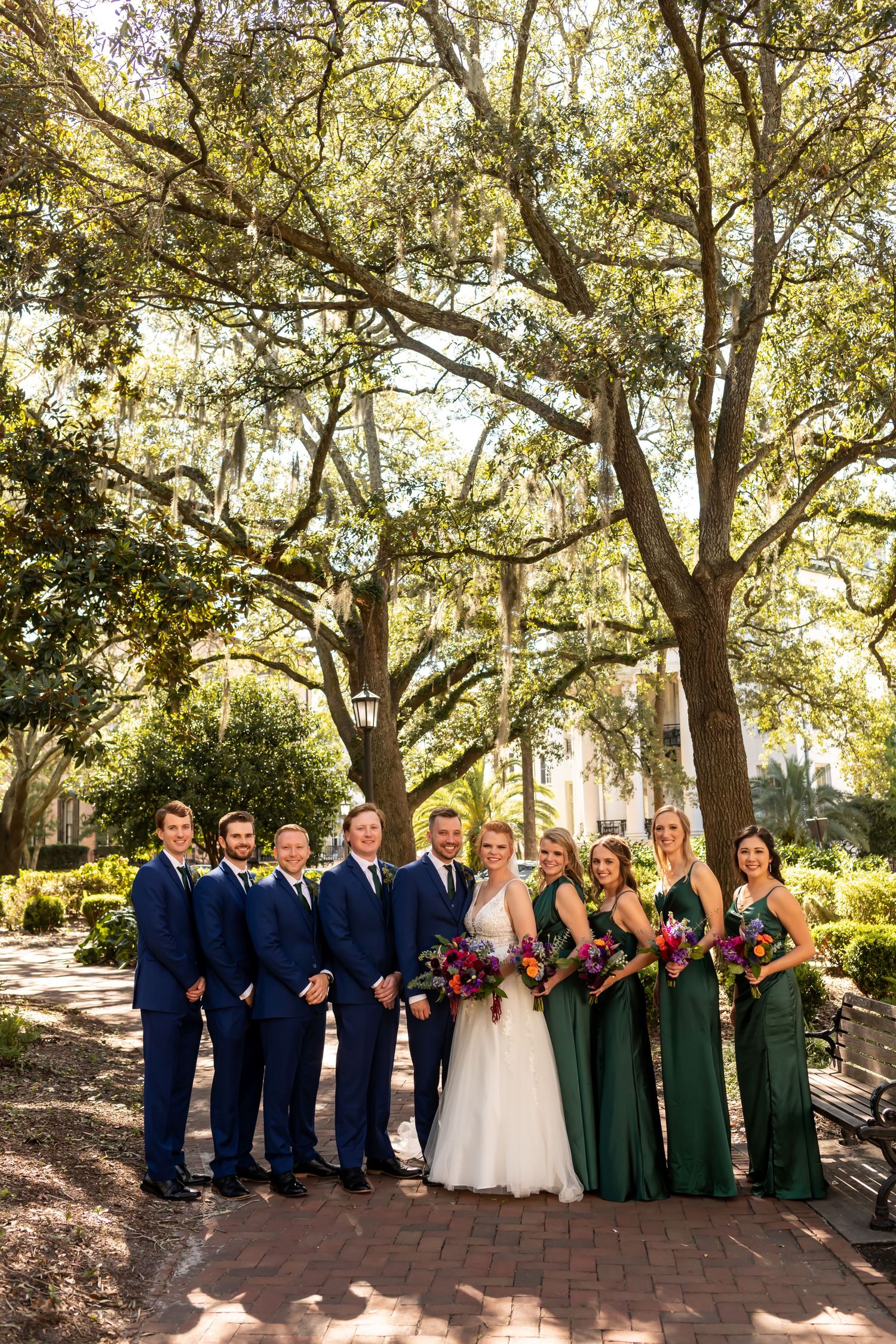 Bridal party poses under Spanish moss draped oak trees on a brick path in a garden setting.