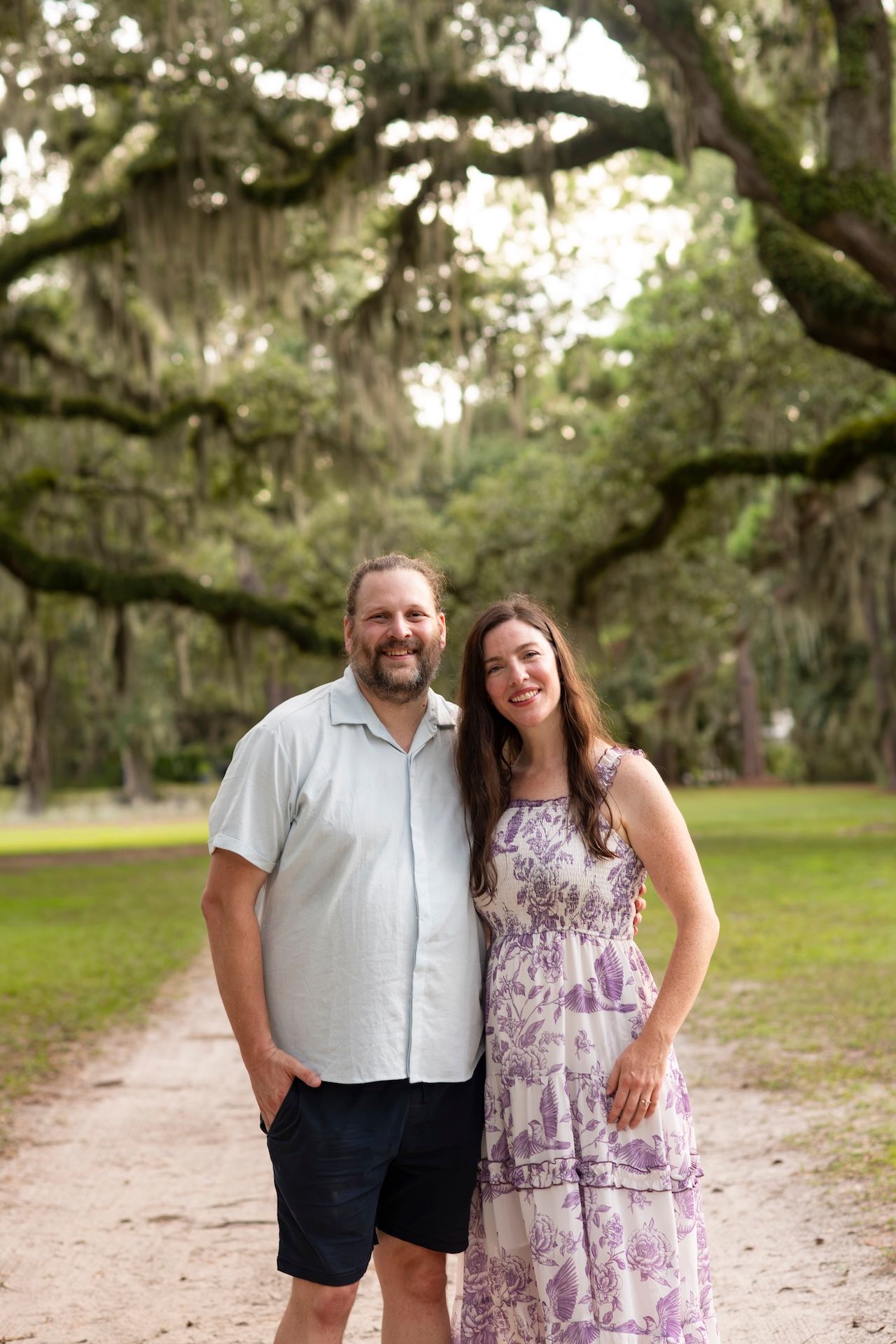 Smiling couple posing on a sandy path surrounded by moss-draped oak trees in a lush green park.