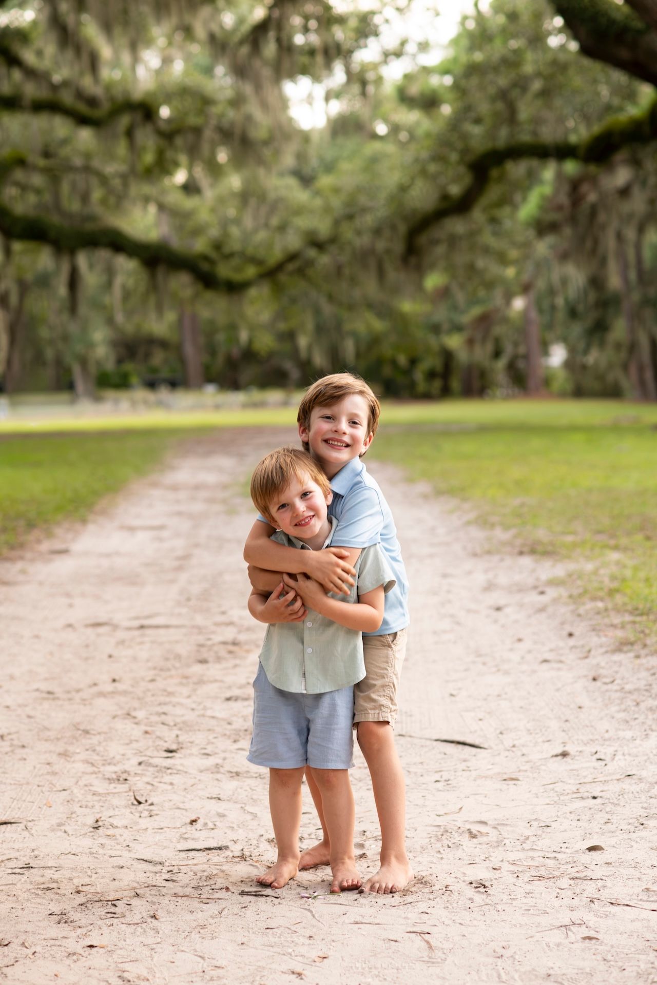 Two young brothers hugging barefoot on a sandy path lined with moss-draped oak trees in a park.
