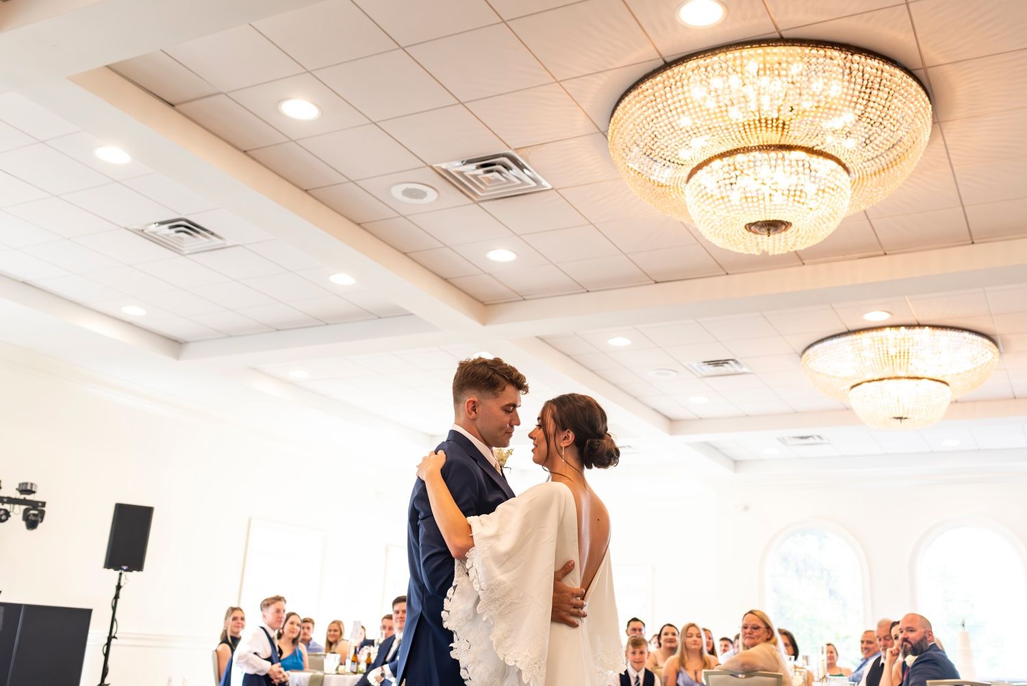 A couple shares their first dance as newlyweds in a grand ballroom with ornate crystal chandeliers overhead.
