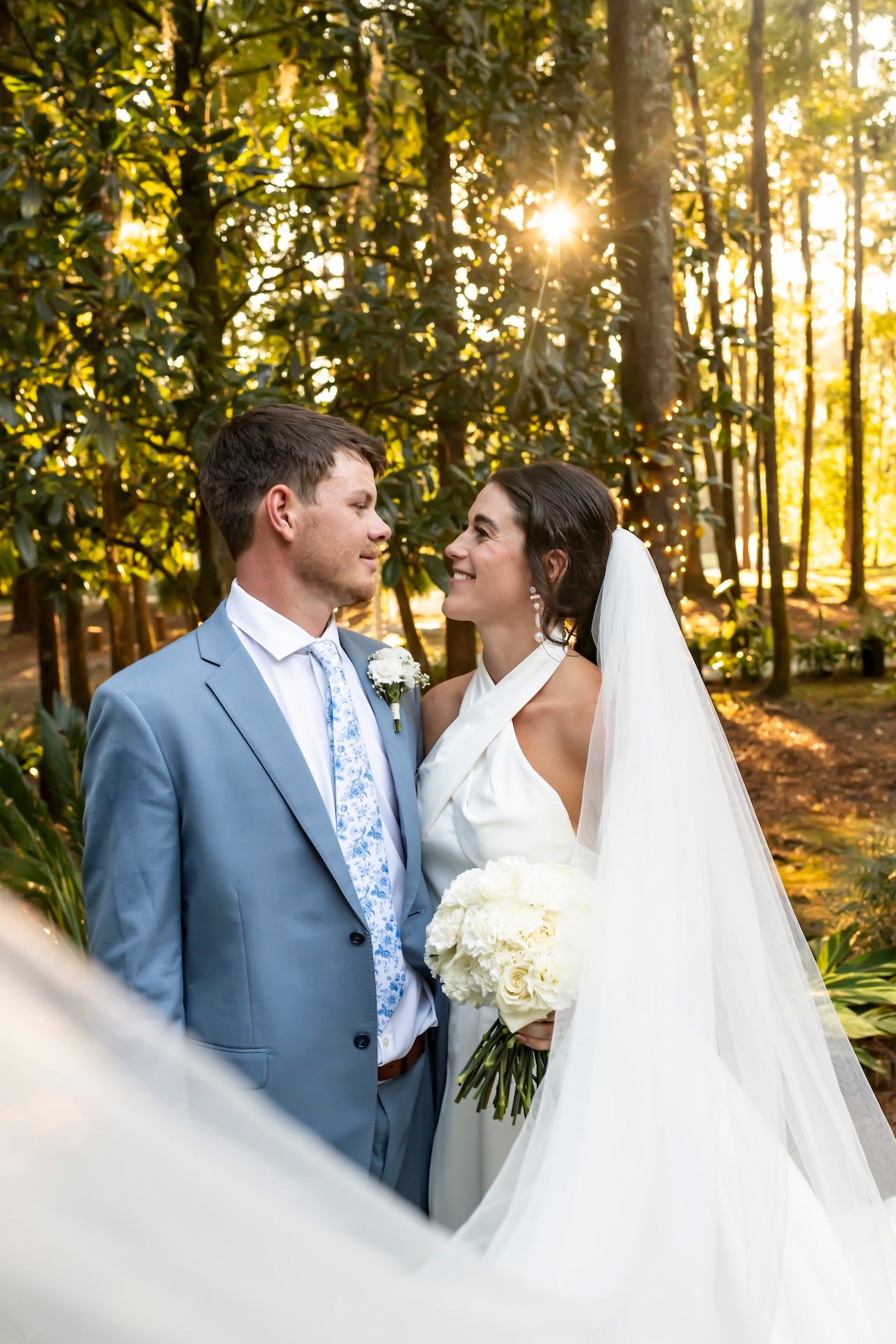 A bride and groom share a romantic gaze outdoors, surrounded by sunlit trees, as she holds a white bouquet.