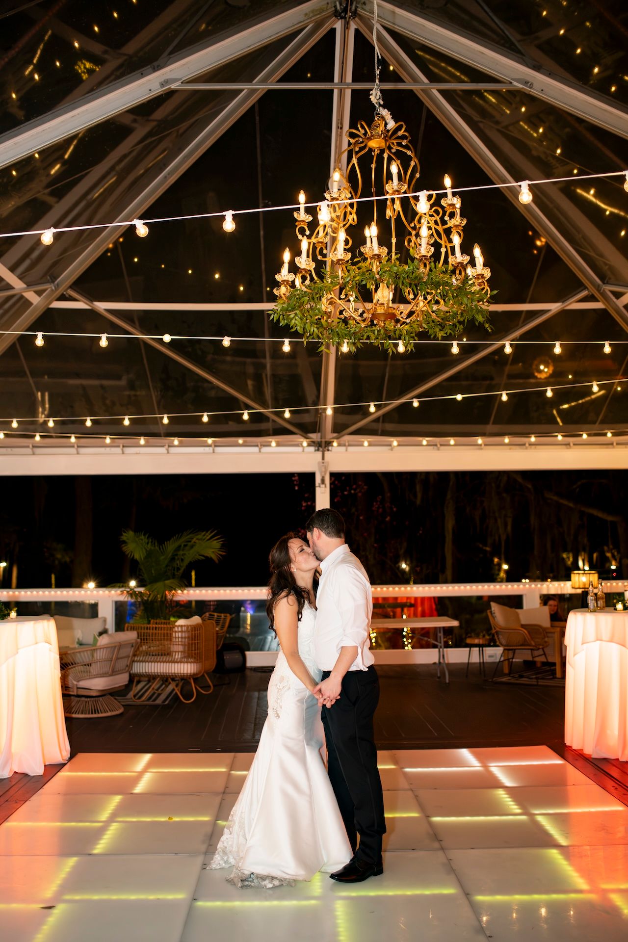 Bride and groom share first dance kiss under chandelier and string lights on illuminated dance floor at night.