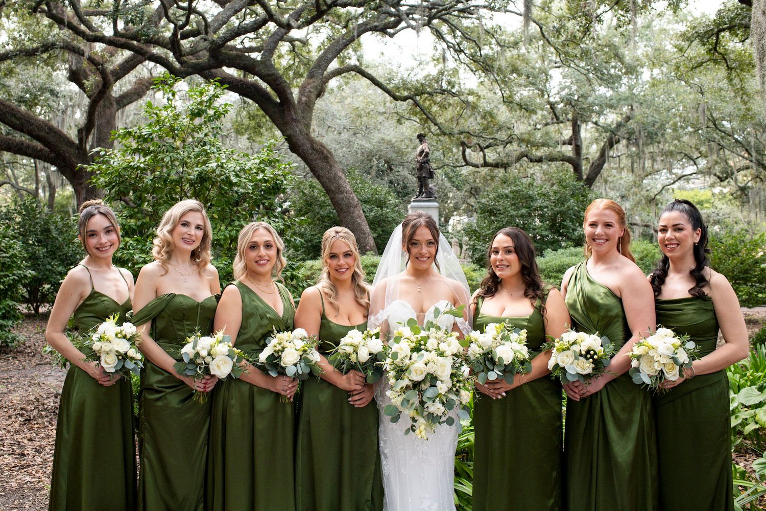 Bridesmaids in olive green dresses pose with bouquets under blooming trees at an outdoor wedding ceremony.