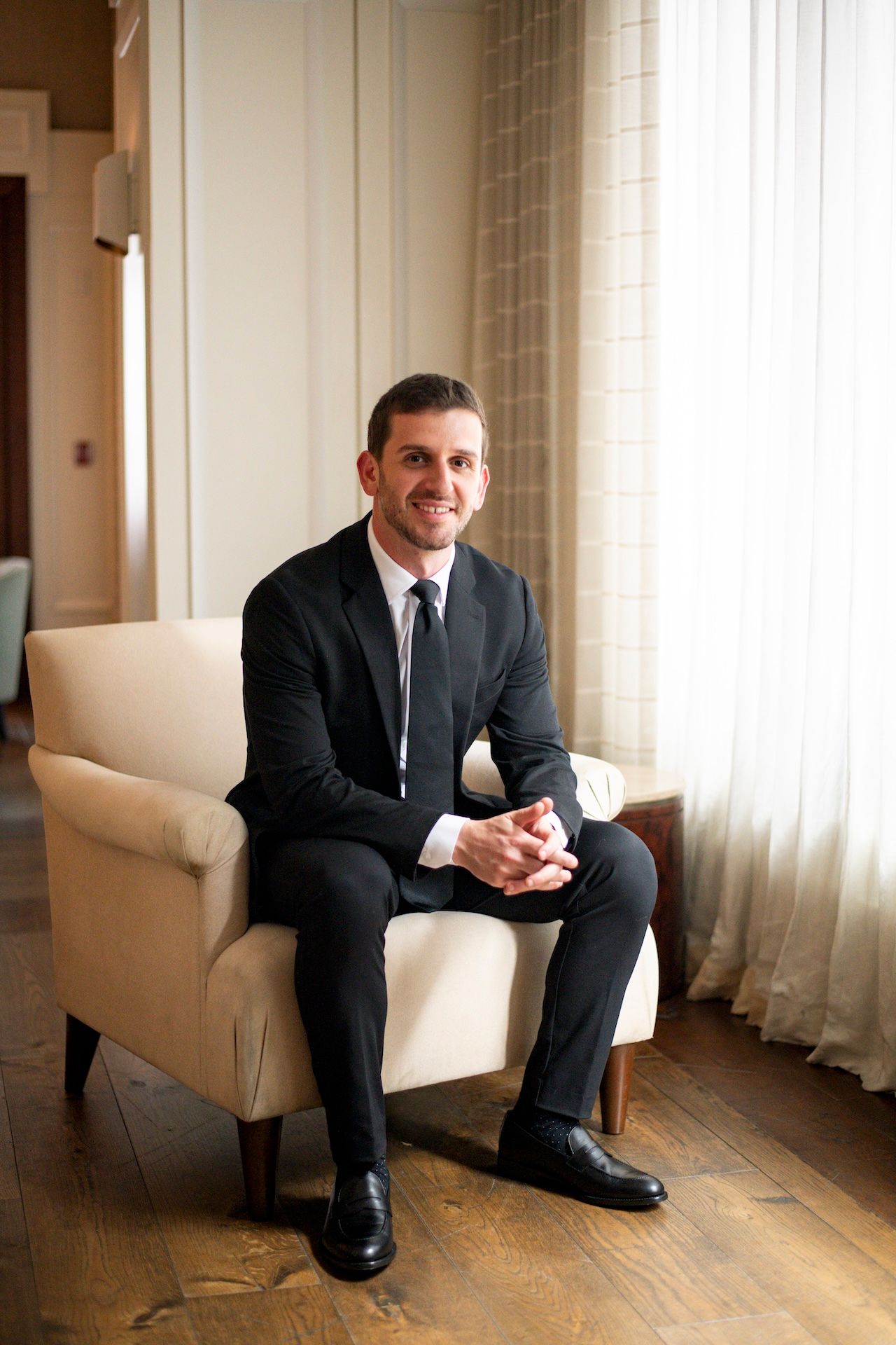 A businessman in a dark suit sits contemplatively in a beige armchair by a sunlit window.