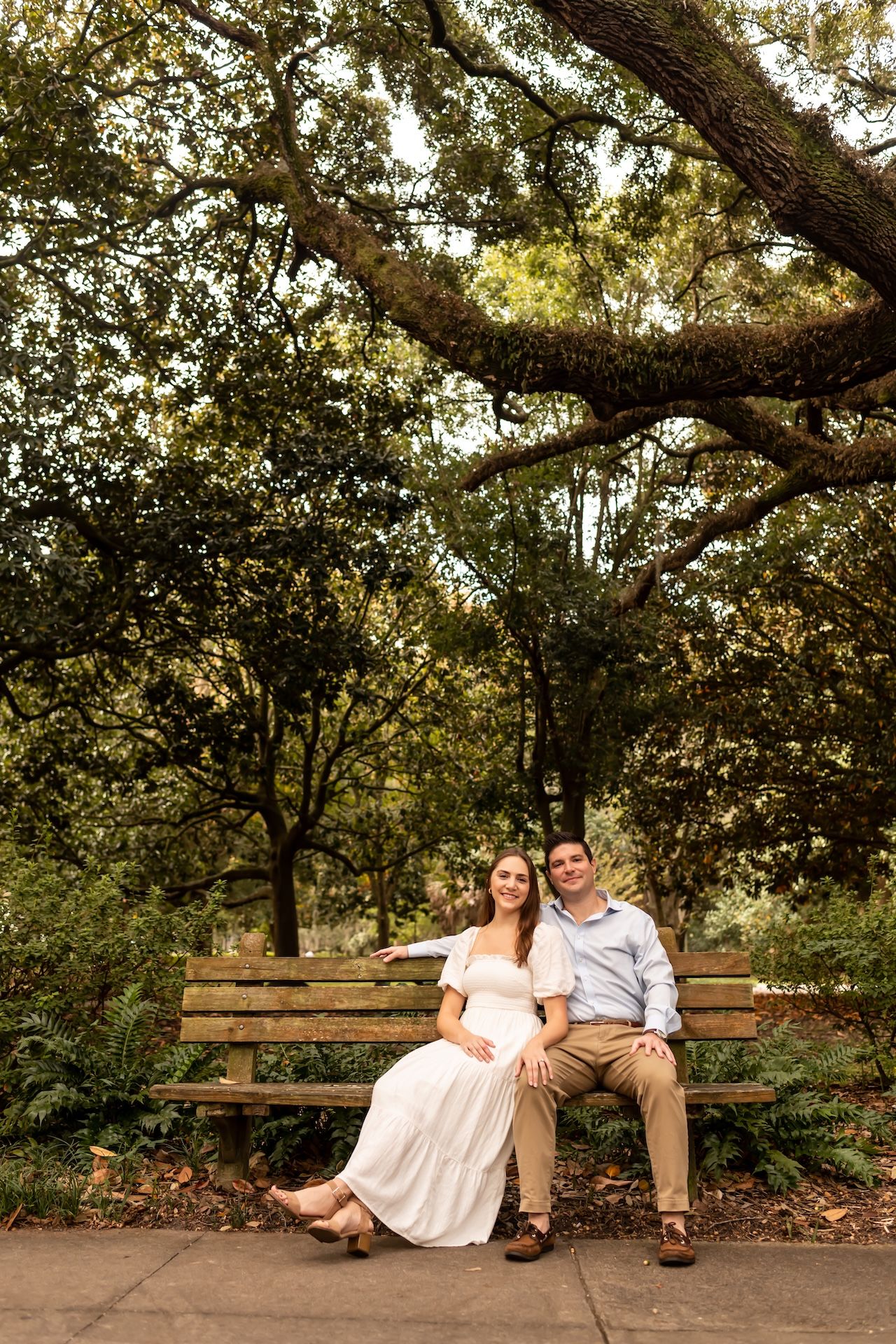 A couple sits together on a wooden bench under lush green trees in a romantic outdoor setting.