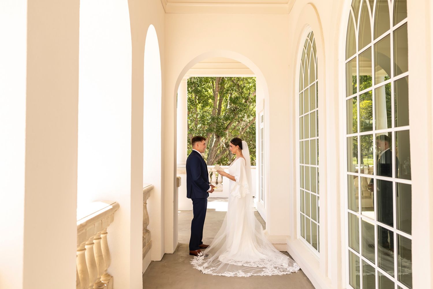 An elegant couple stands under a white columned portico with arched windows and lush greenery in the background.