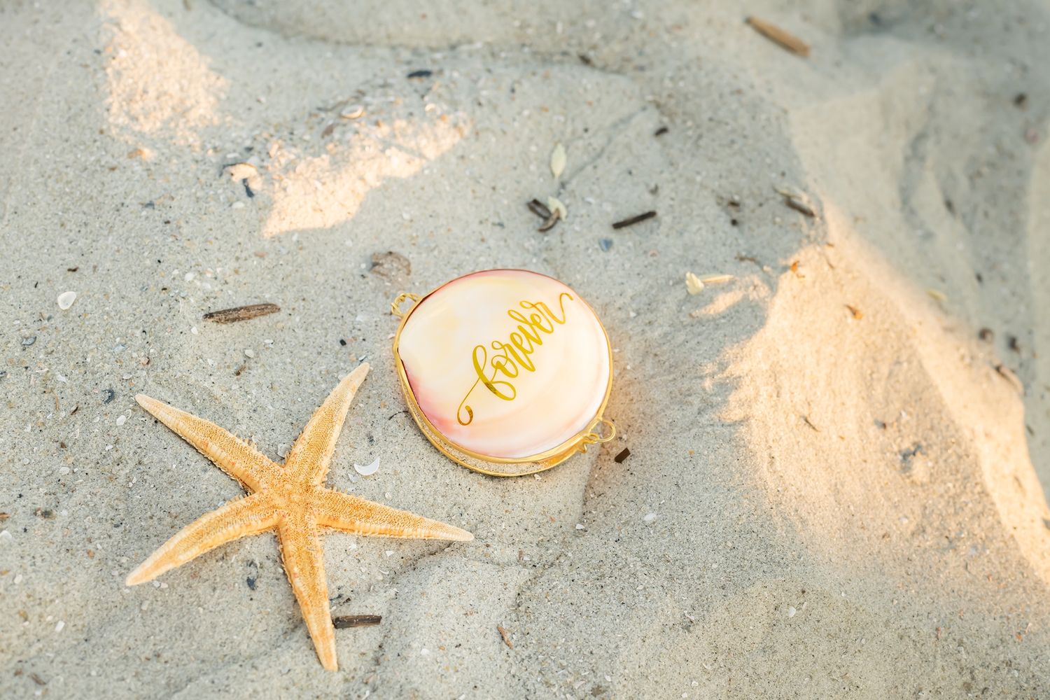 A golden wedding ring and orange starfish rest on soft white beach sand in natural lighting.