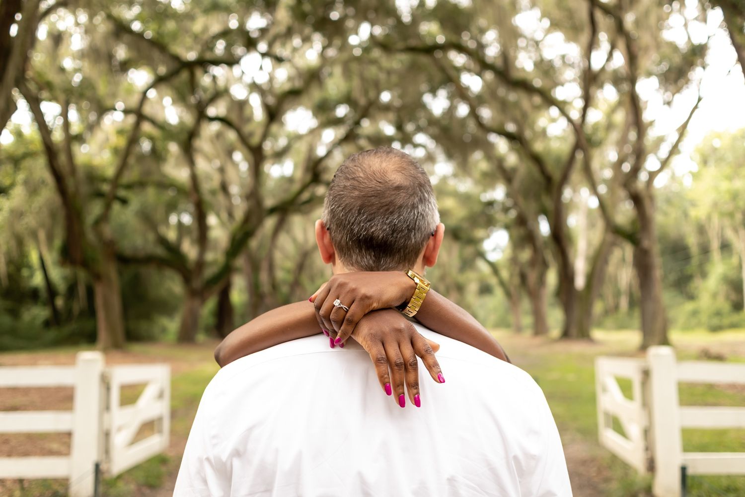 Person in white shirt seen from behind with hands clasped behind neck standing in front of tree-lined path with white fence.