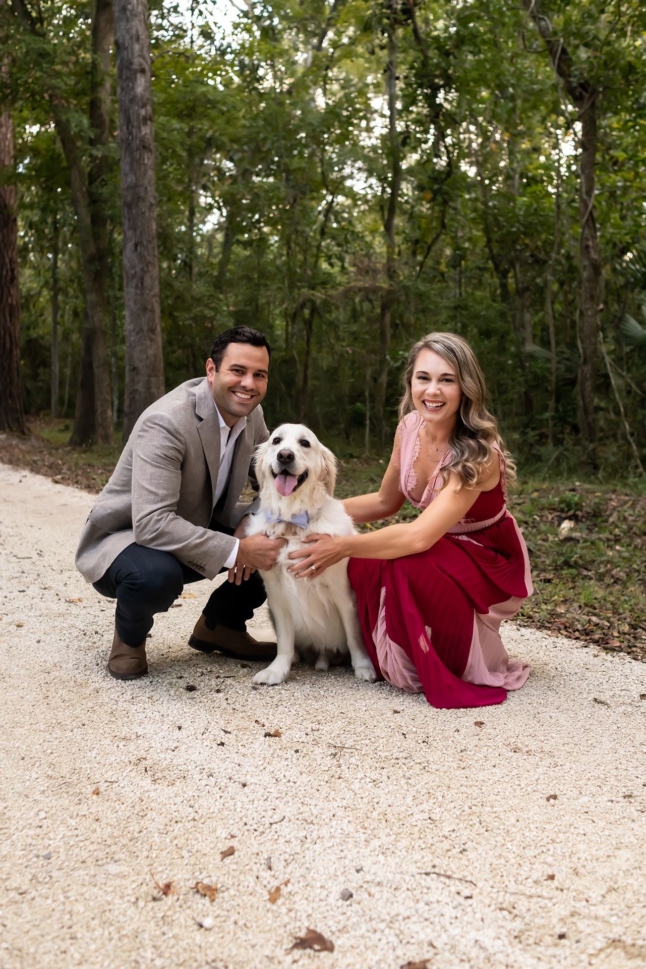 A golden retriever poses with two people on a gravel path in a wooded area.