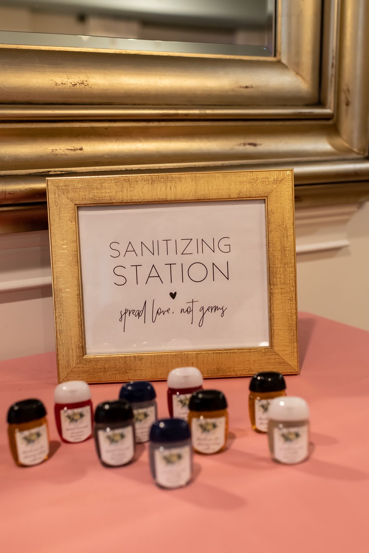 A row of small glass jars with labels sits under a wooden-framed 'Sanitizing Station' sign on a pink surface.
