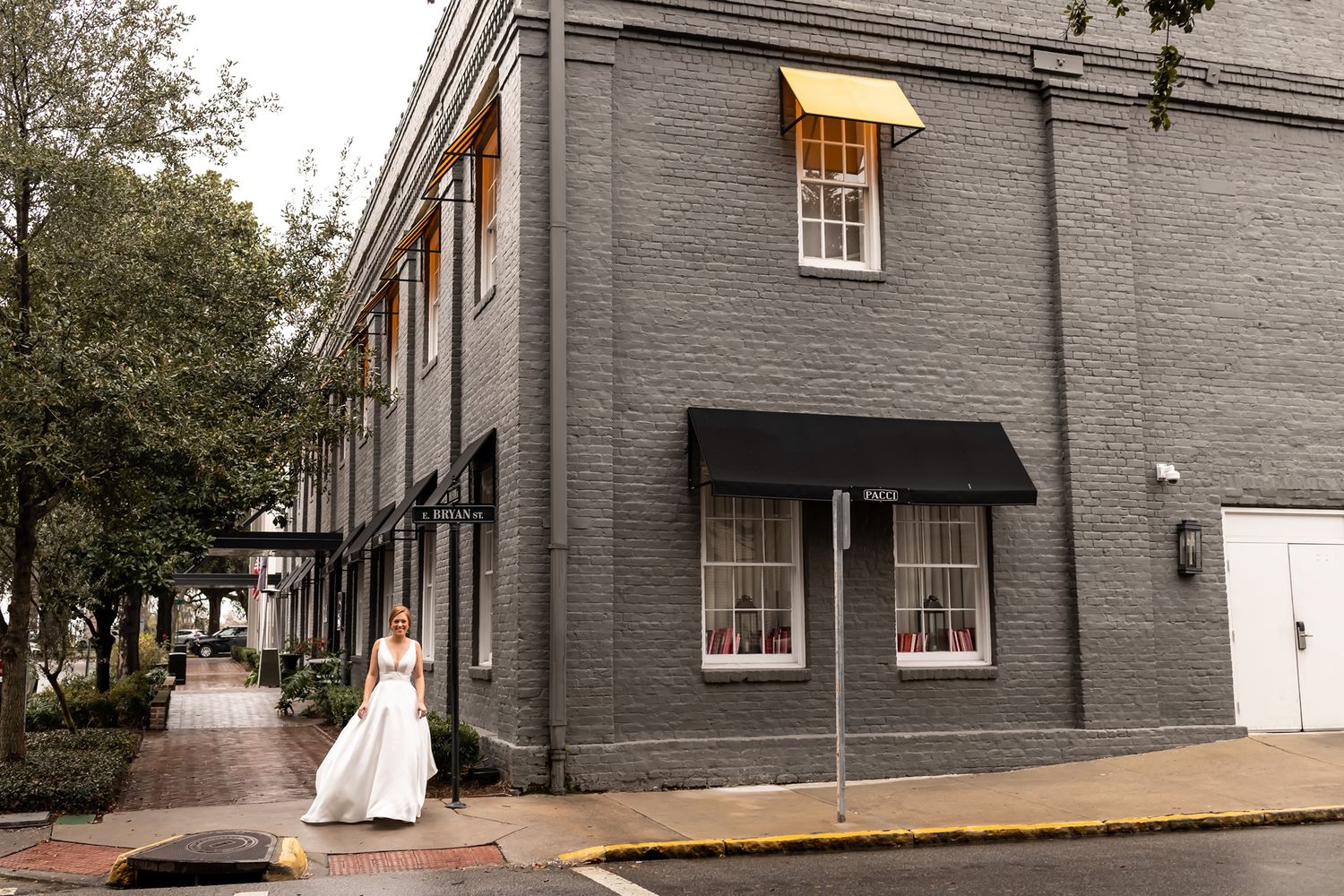 A figure in a white gown walks past a gray brick building with black awnings and windows on a historic street corner.