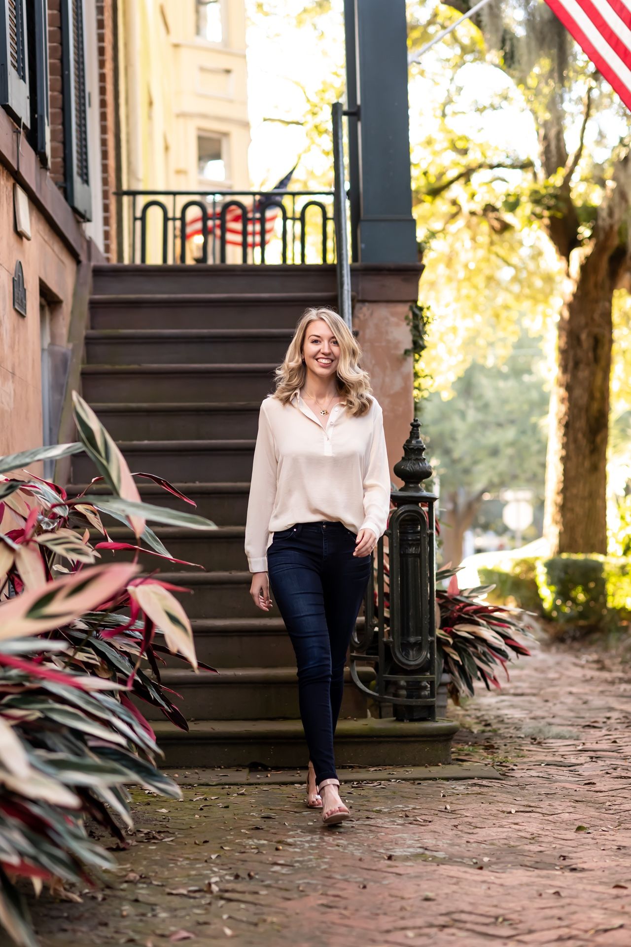 A person in a beige sweater and jeans walks along a brick-lined path with American flags and foliage.