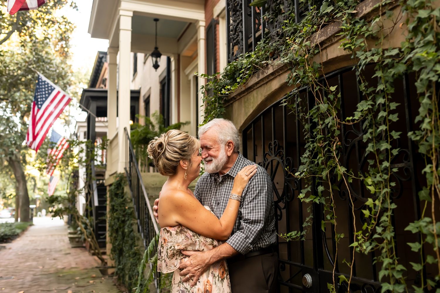 A warm embrace shared between two people on a charming brick sidewalk with American flags and greenery in the background.