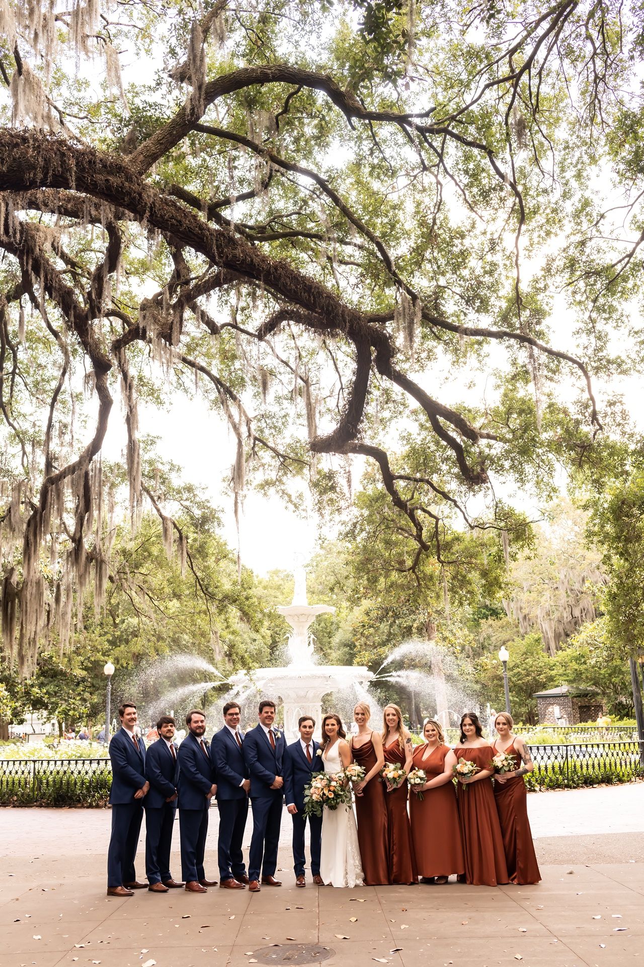 Wedding party poses under moss-draped oak trees near a fountain in a scenic southern garden setting.