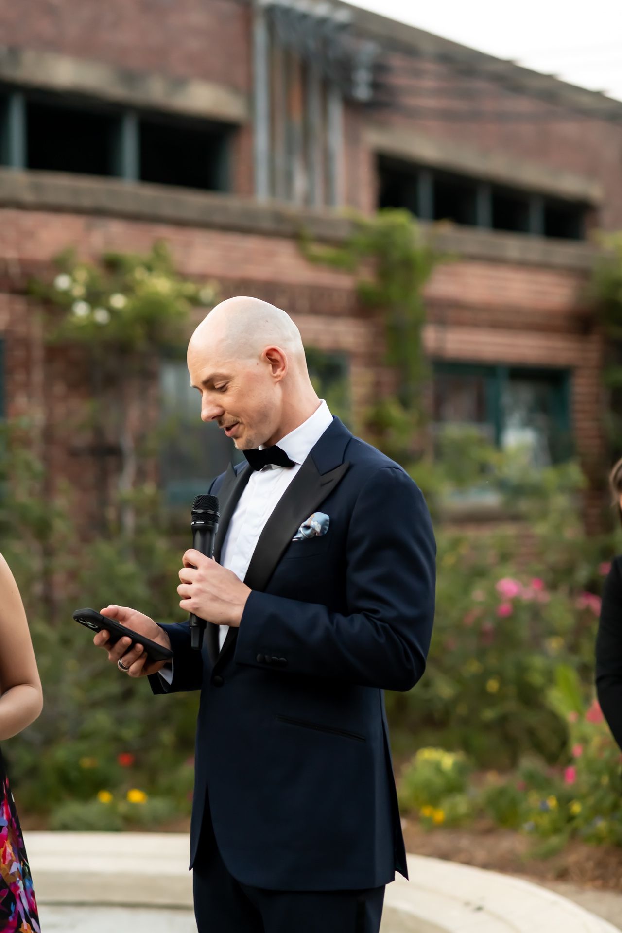 A person in a black tuxedo and bow tie consults their phone while holding a microphone at an outdoor event.