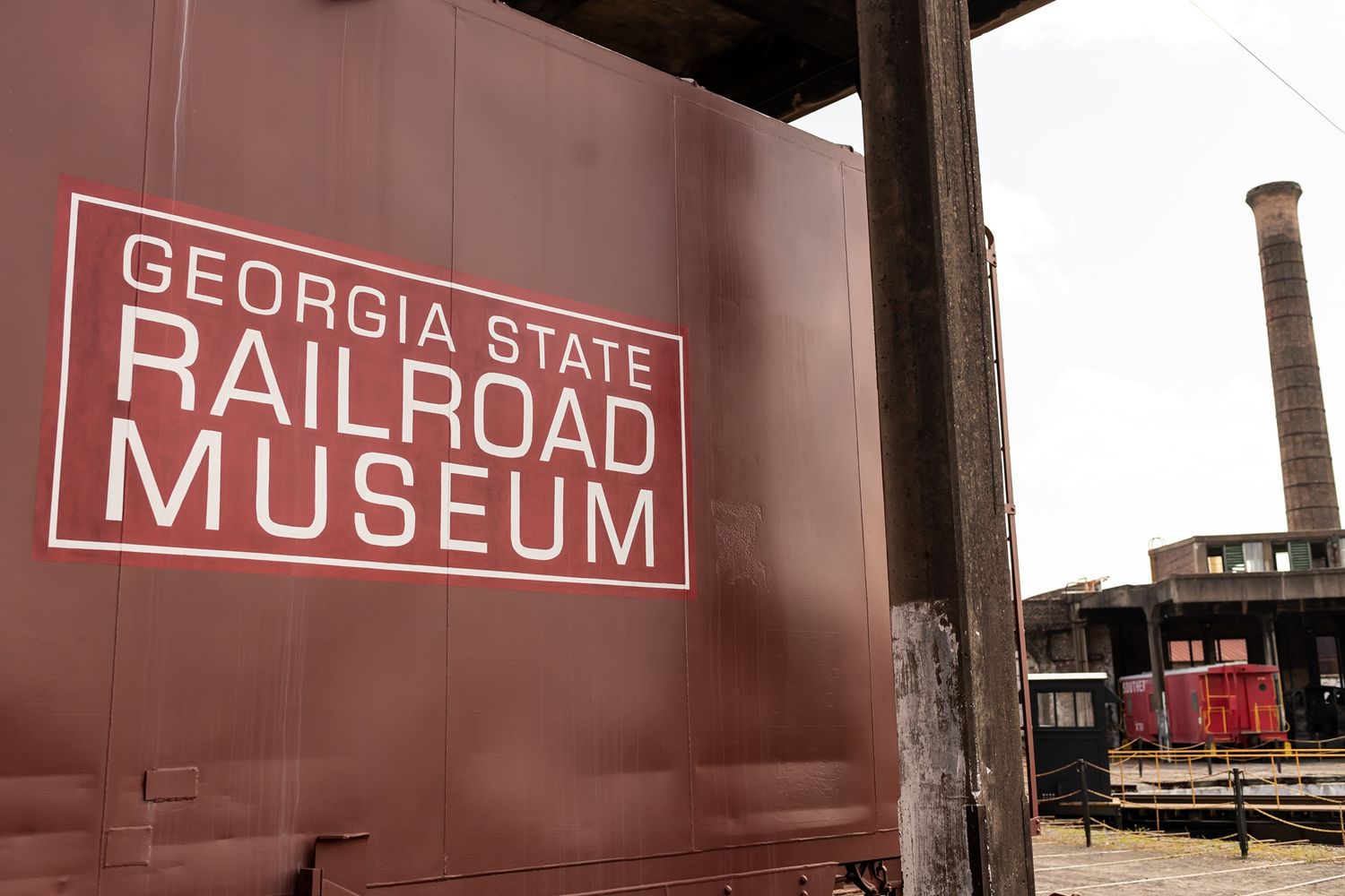 Red sign for Georgia State Railroad Museum mounted on rustic wall with vintage train car and smokestack visible in background.