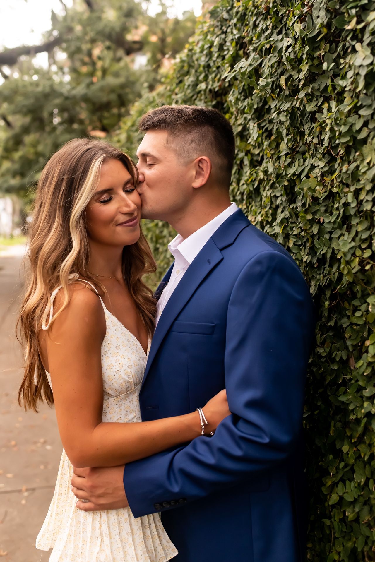 Romantic couple embraces against green hedge wall outdoors with warm lighting in elegant wedding attire.
