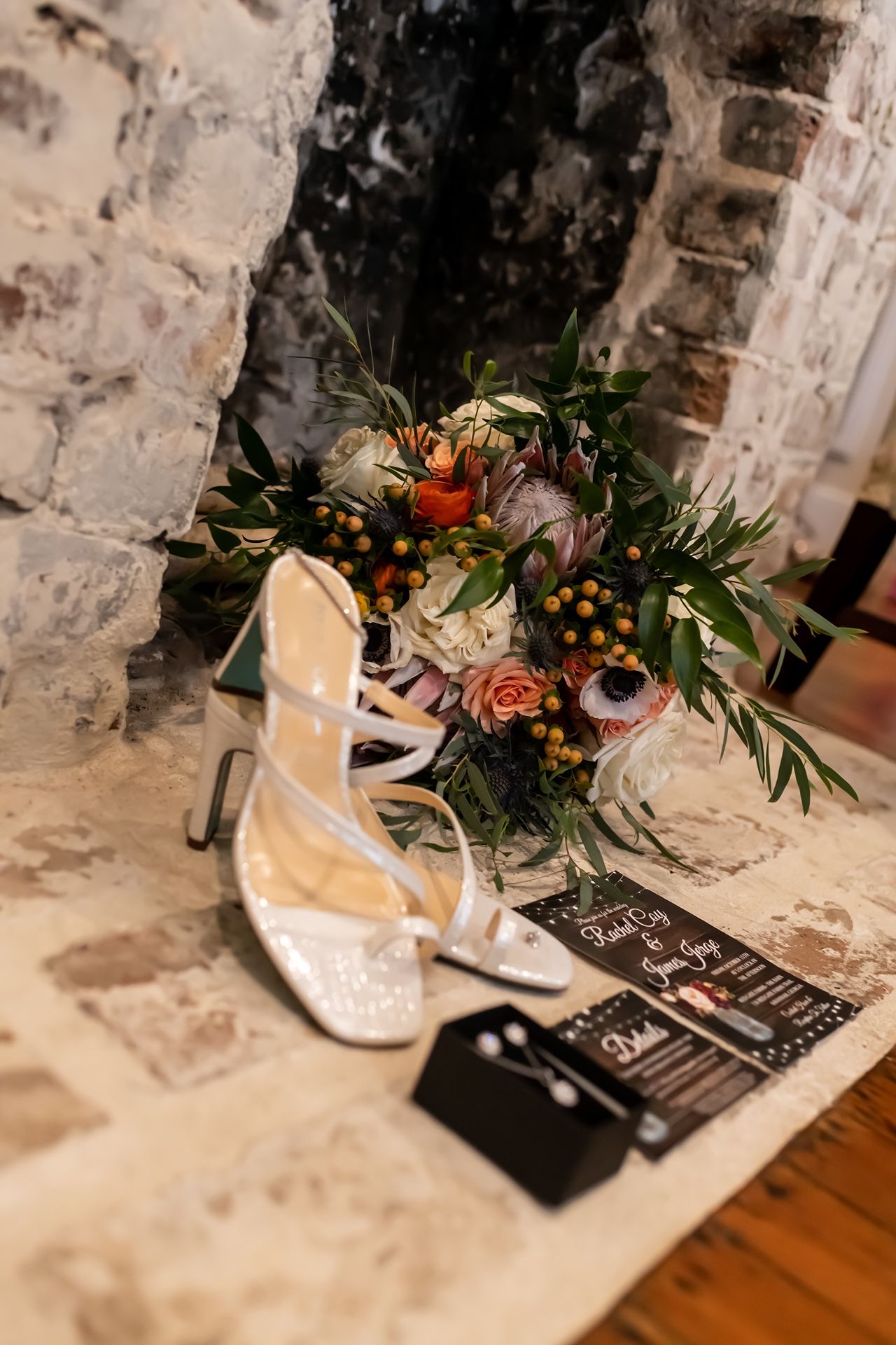 White bridal shoes and delicate floral bouquet with roses and greenery arranged next to stone wall and wooden floor.