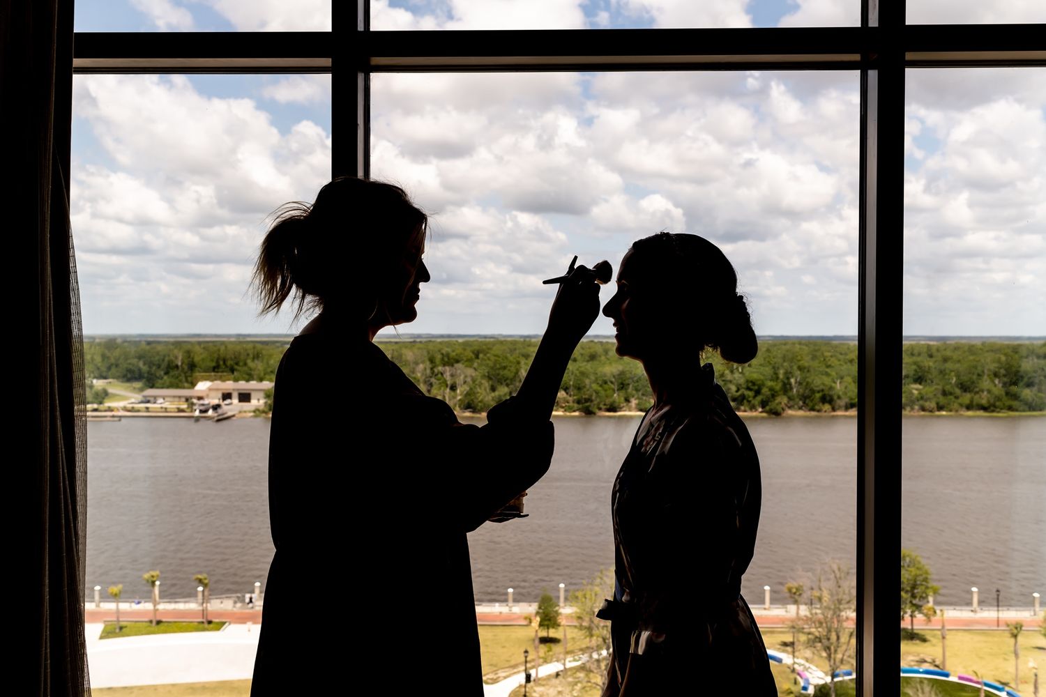 Silhouette of two figures against a window with water view, one applying makeup to the other.