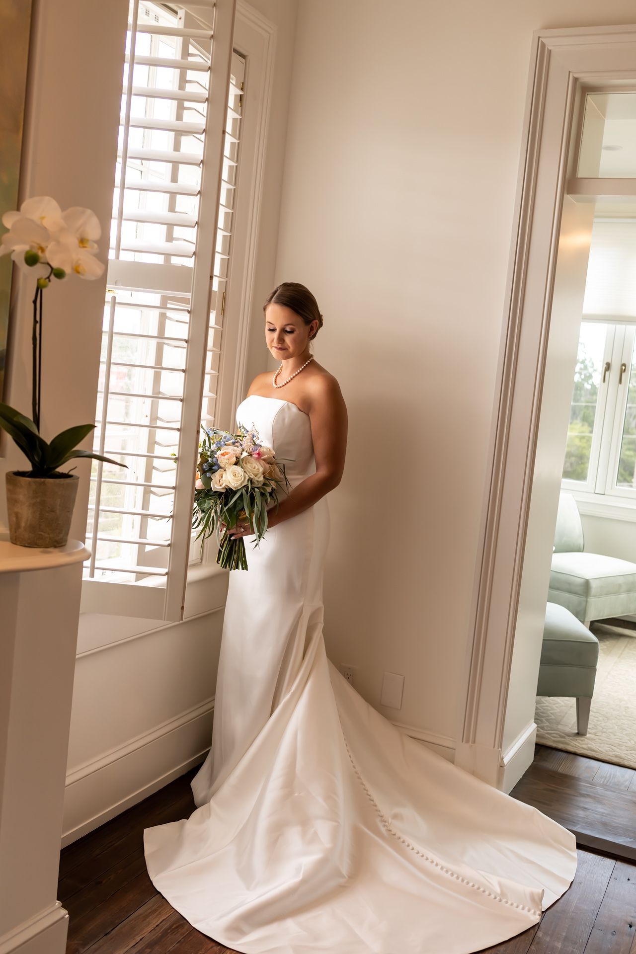Someone in an elegant white wedding dress stands in a bright room with white shuttered windows and an orchid plant.