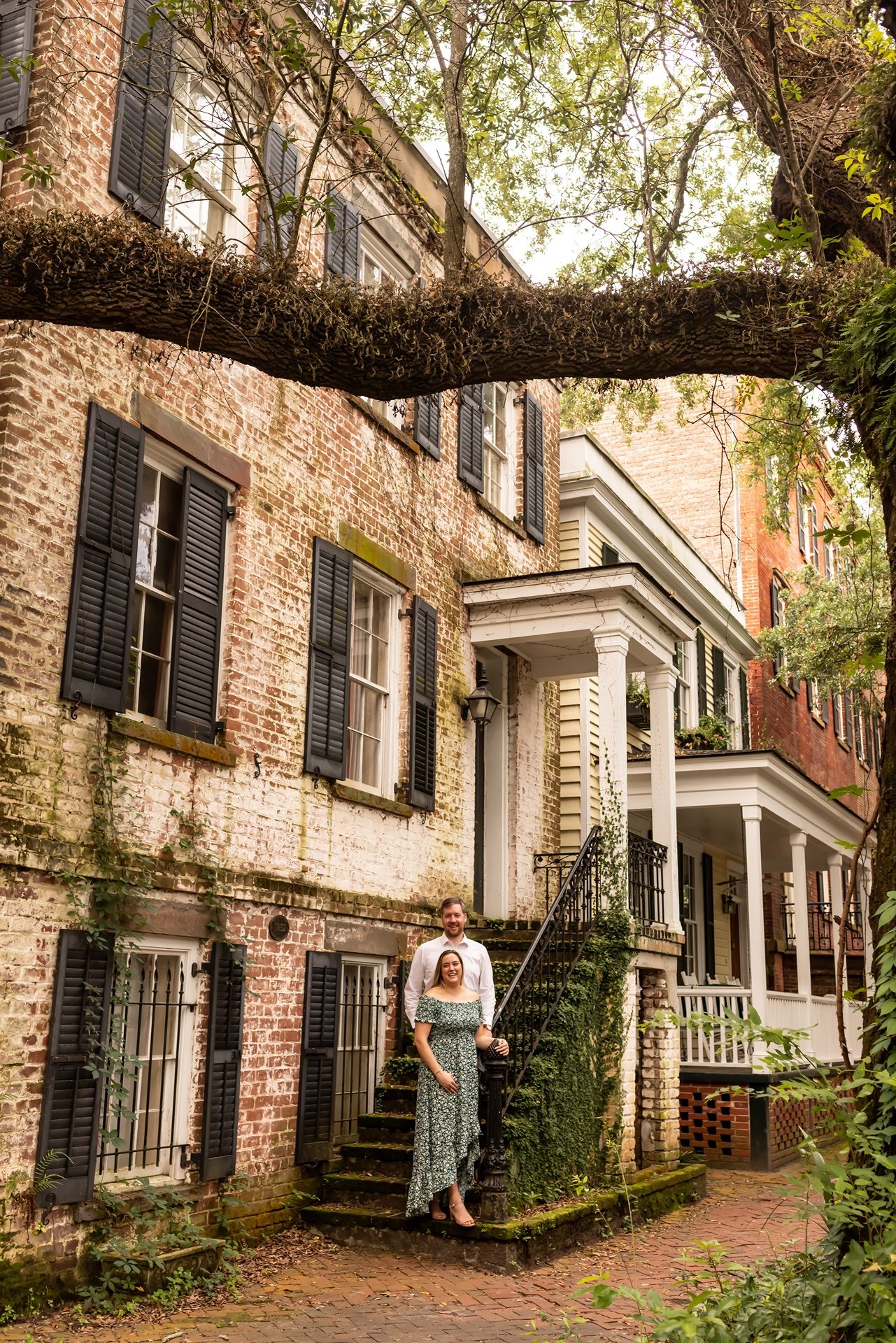 Moss-covered brick pathway leading to historic townhouses with black shutters and white columns.