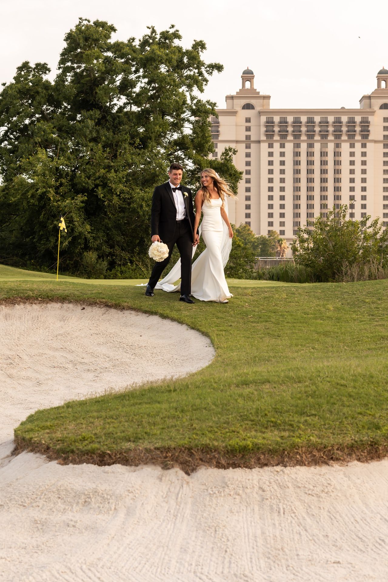 A couple walking on a golf course with sand bunkers and a luxury resort hotel building in the background.