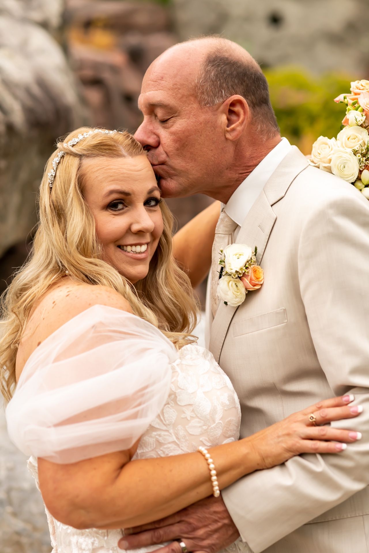 A romantic wedding portrait shows a couple embracing outdoors, with the groom in beige suit kissing his smiling bride.
