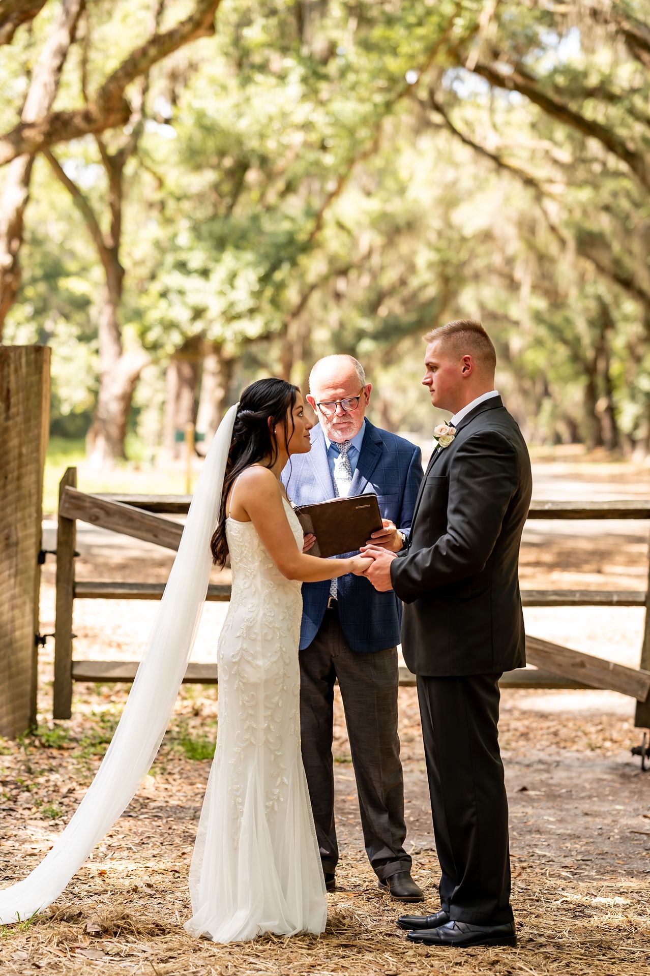 A couple exchanges vows during an outdoor wedding ceremony under moss-draped trees while an officiant presides.