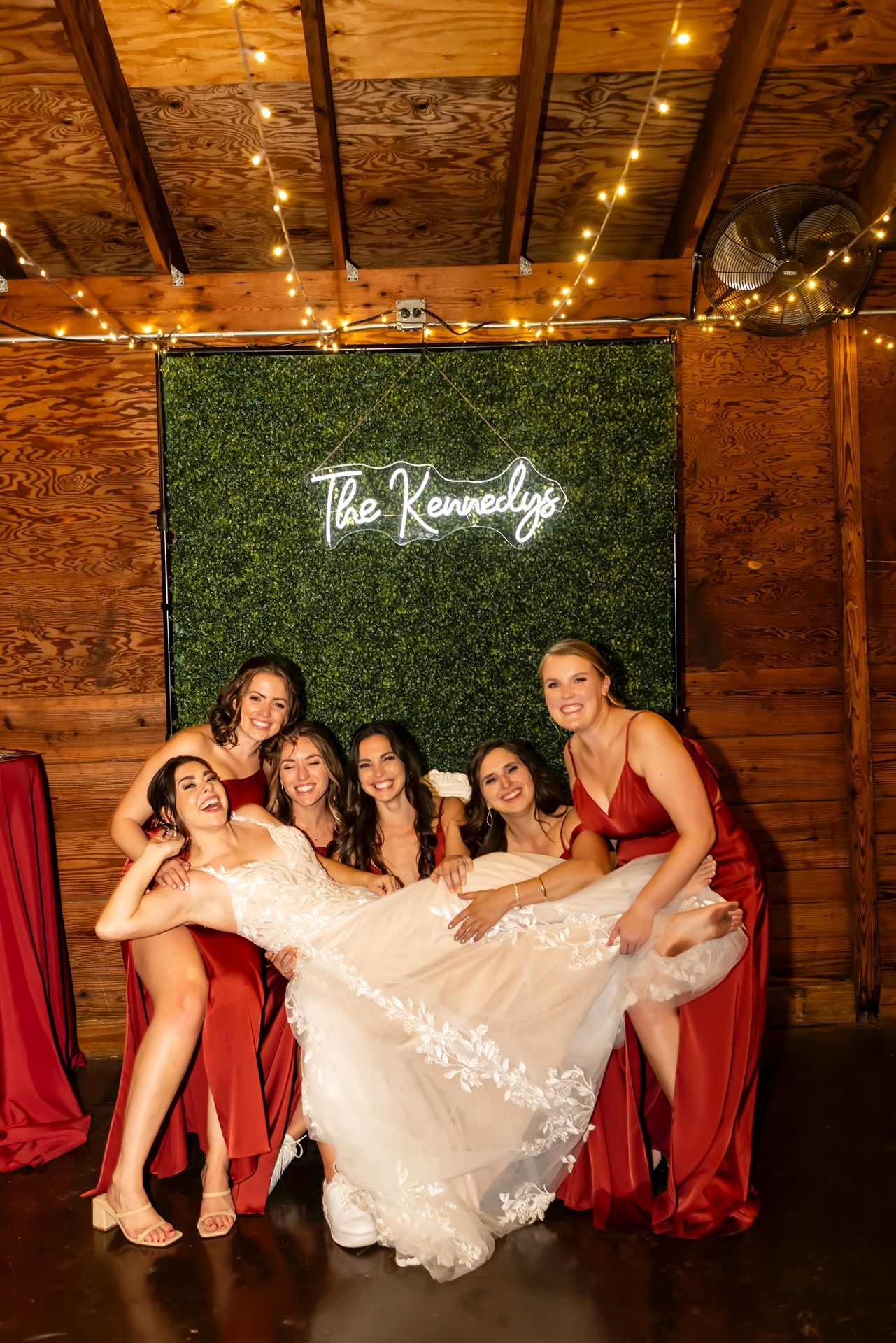 Bride in white dress poses with bridesmaids wearing red dresses in front of illuminated hedge wall with neon sign.