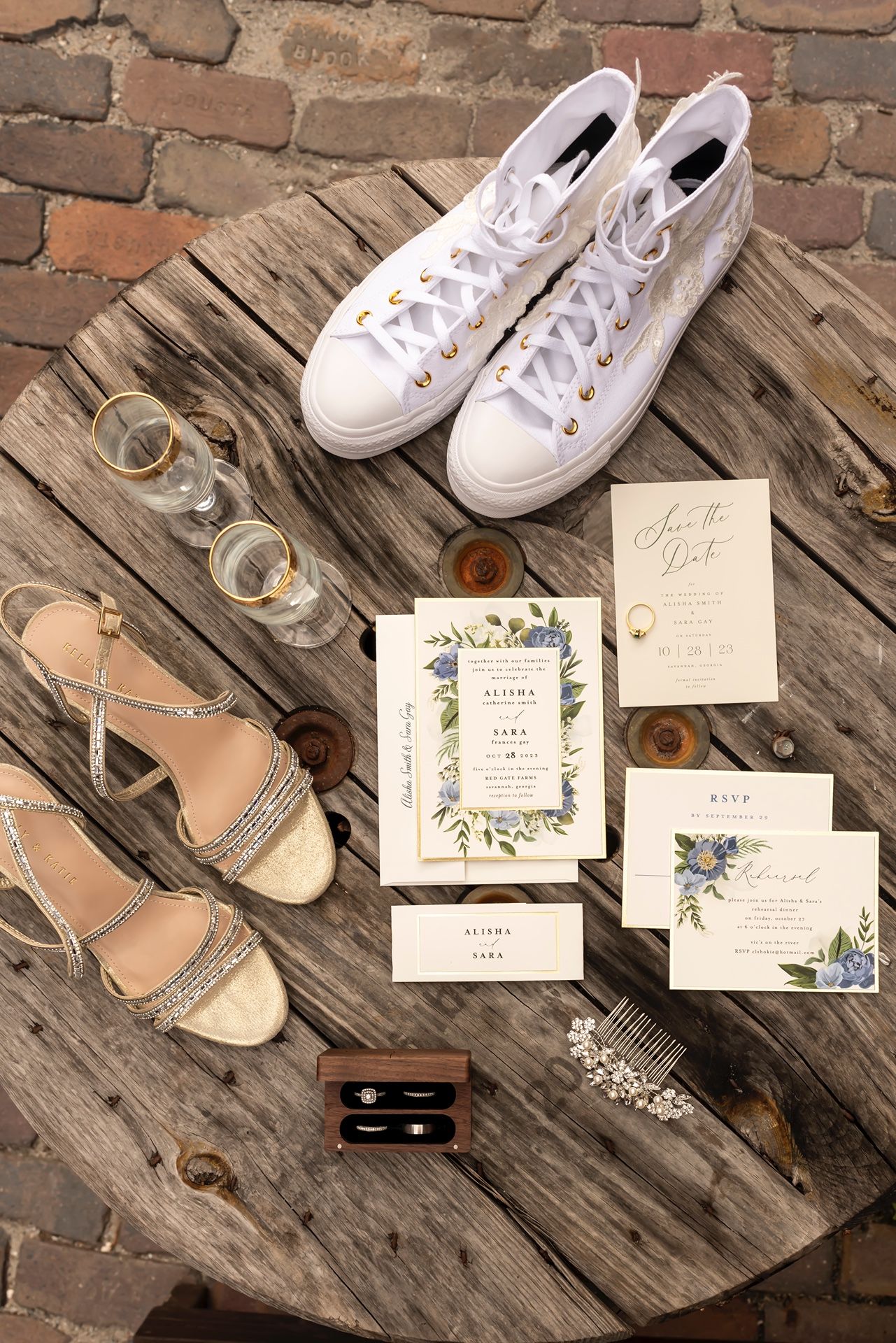 Wedding accessories including white Converse sneakers, silver sandals, floral invitations and glassware on rustic wooden surface.