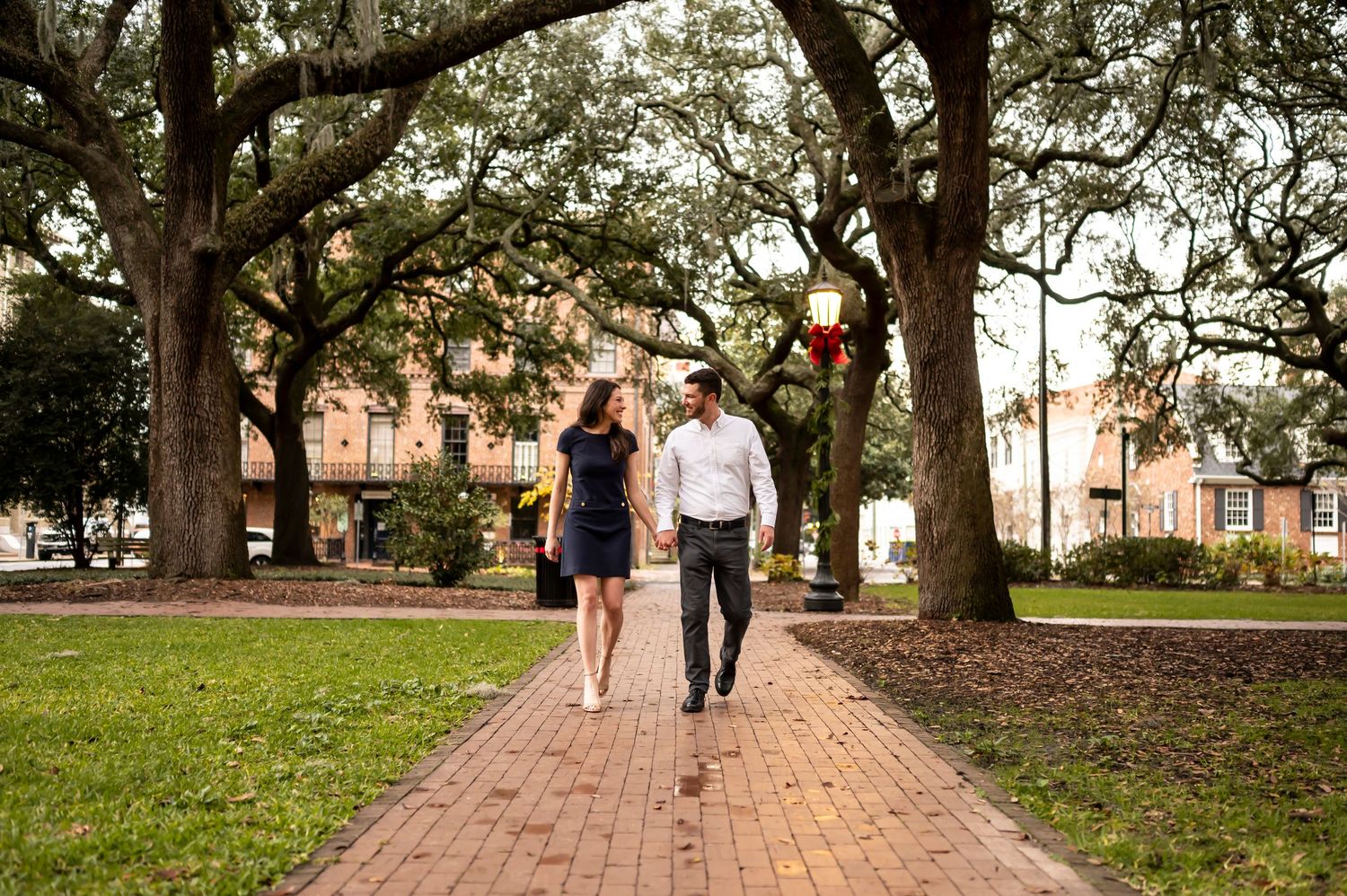 A couple strolls down a brick path lined with majestic oak trees in a scenic park during golden hour.