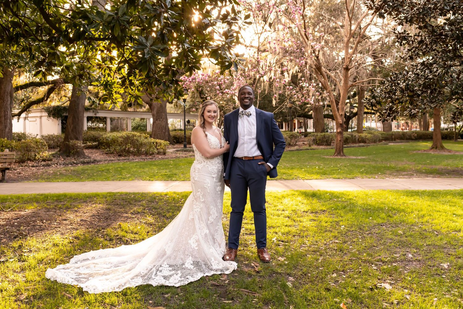 A bride in a lace wedding gown and groom in a navy suit pose together in a sunlit garden park setting.