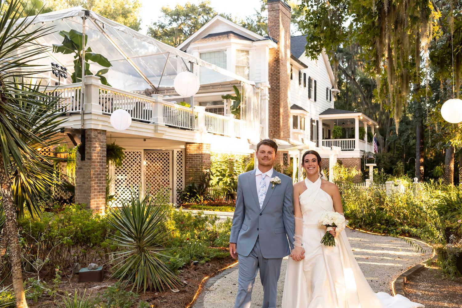 Wedding couple poses in front of historic buildings with sunlit trees and landscaping in the background.