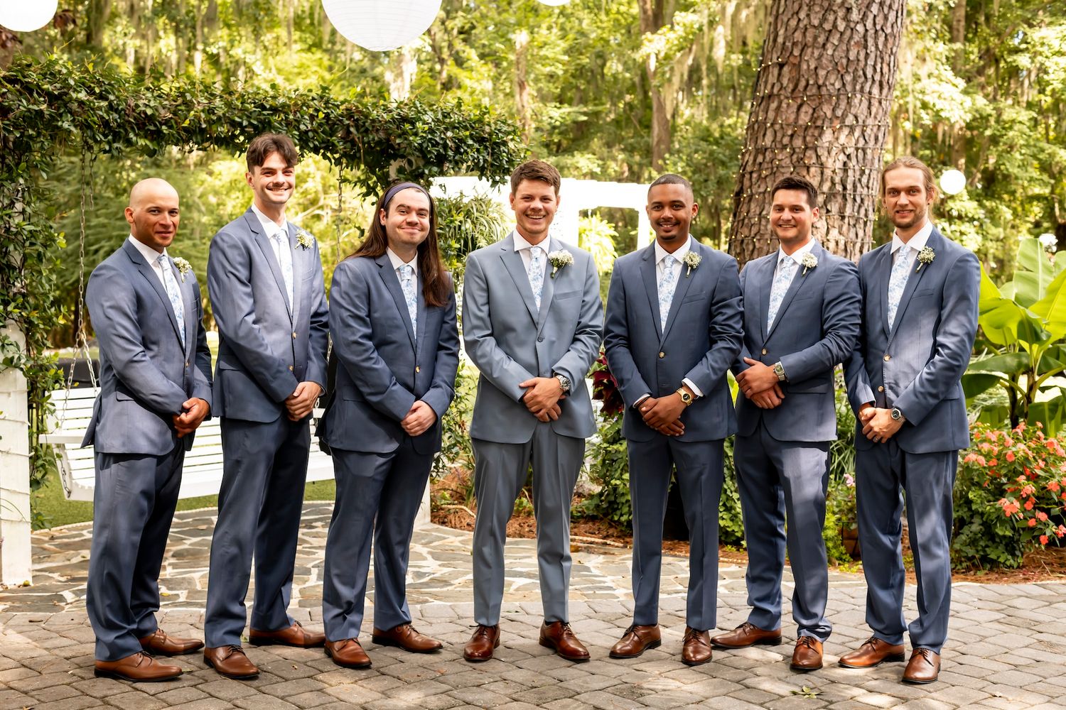 Groomsmen in matching gray suits and brown shoes stand together in a garden setting for a wedding photo.