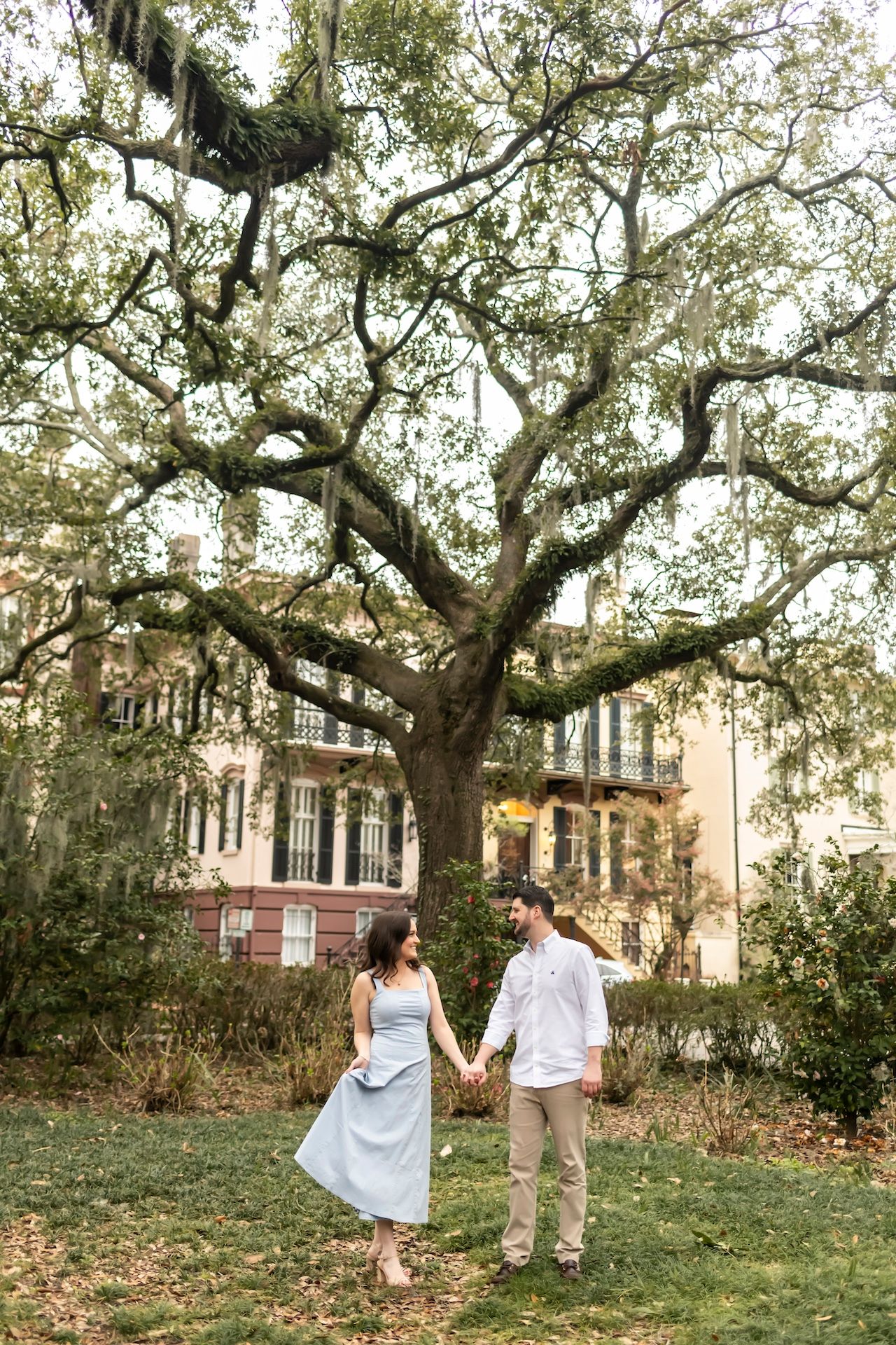 A couple walks hand in hand beneath a grand oak tree draped in Spanish moss outside a historic southern mansion.