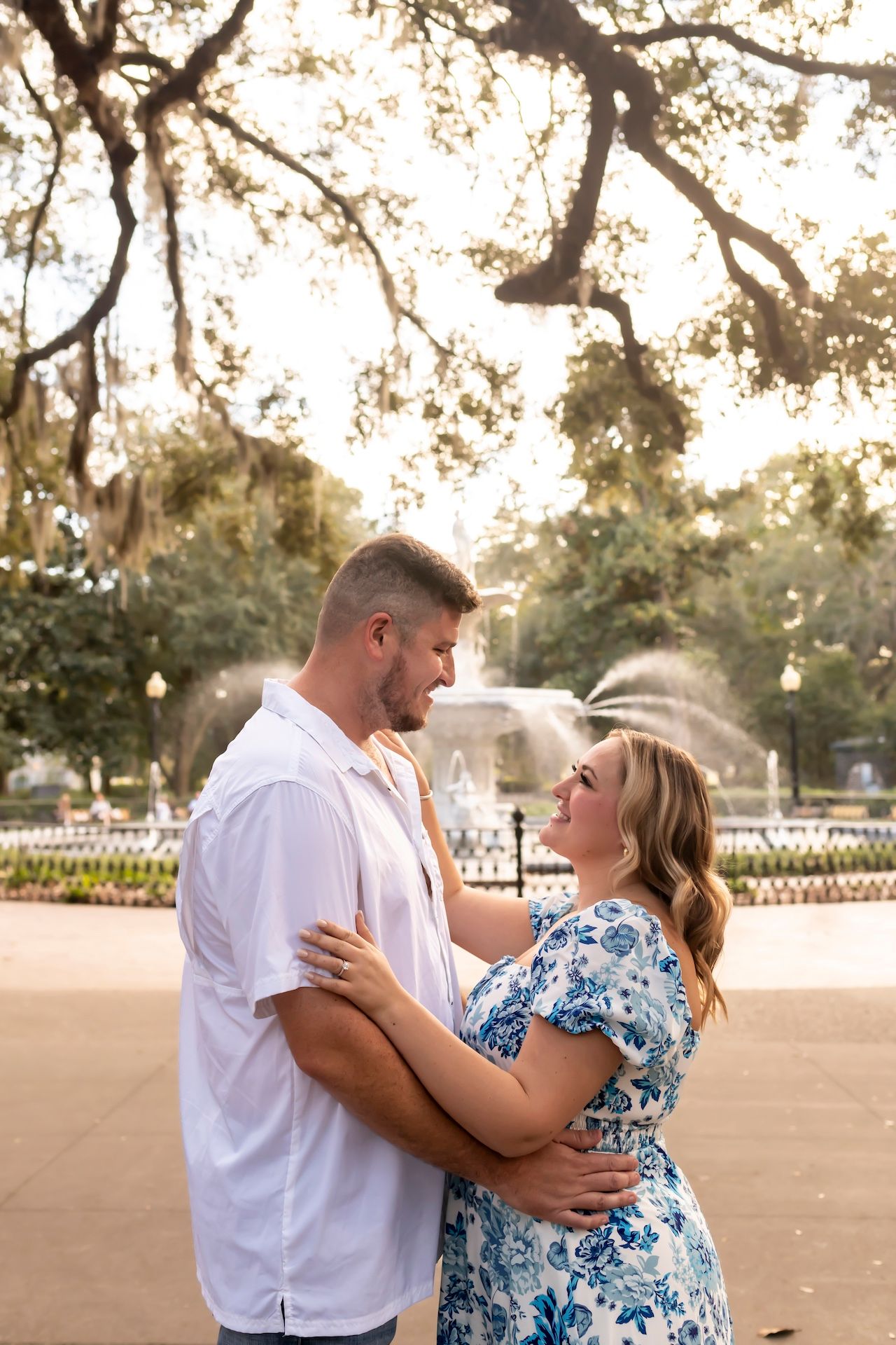 A couple embraces outdoors at sunset under tree branches at a park in their casual summer attire.