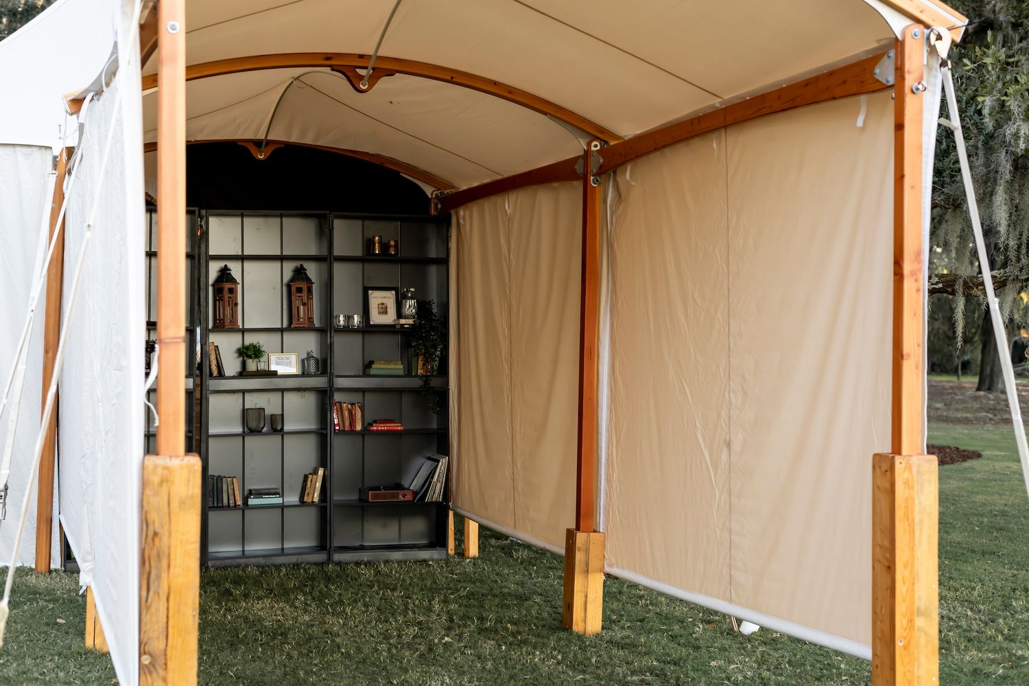 A rustic canvas tent with wooden poles houses bookshelves filled with books, creating a temporary outdoor library.