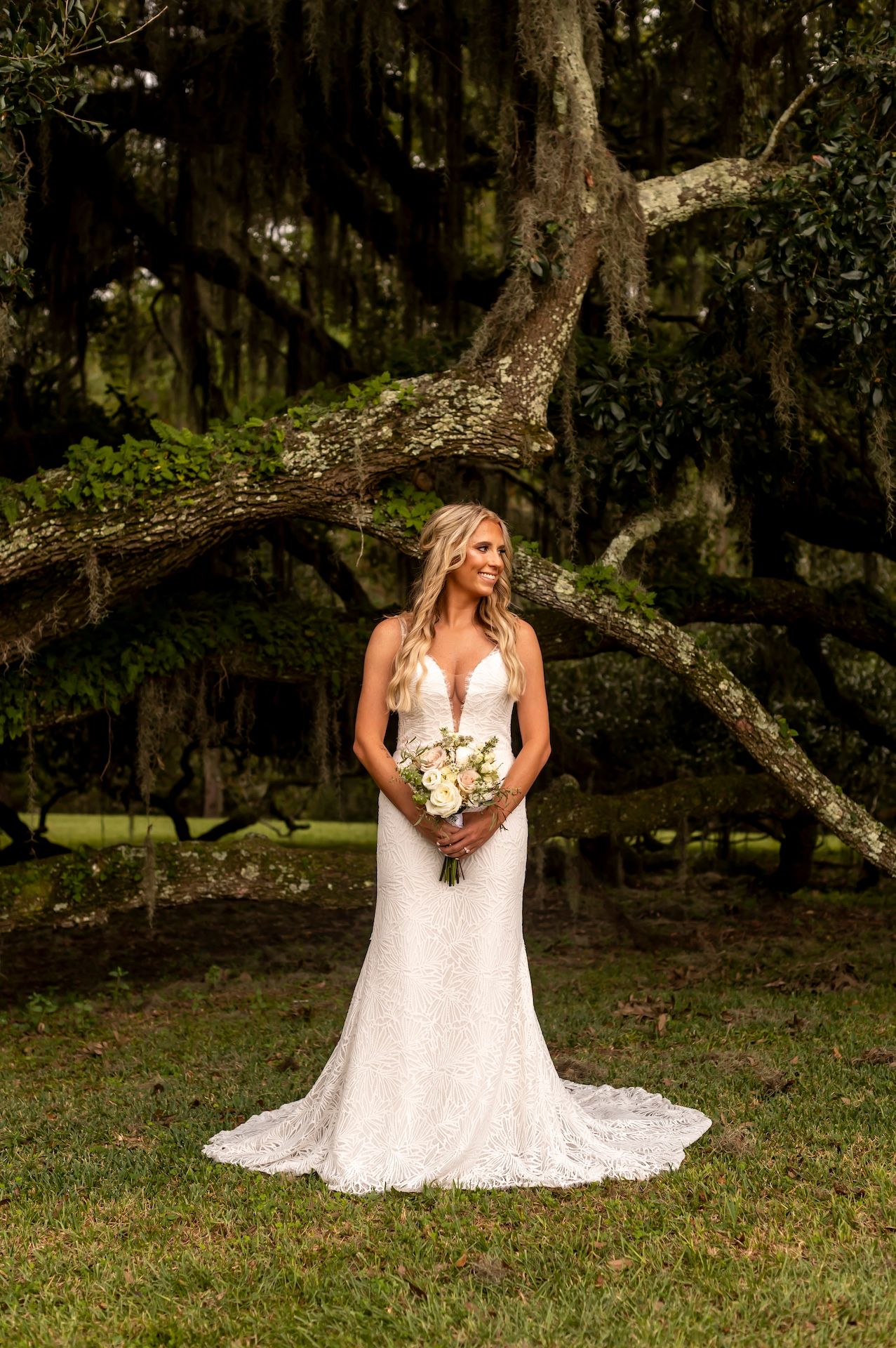 A stunning white lace wedding gown with a train photographed under a mossy oak tree in a natural outdoor setting.