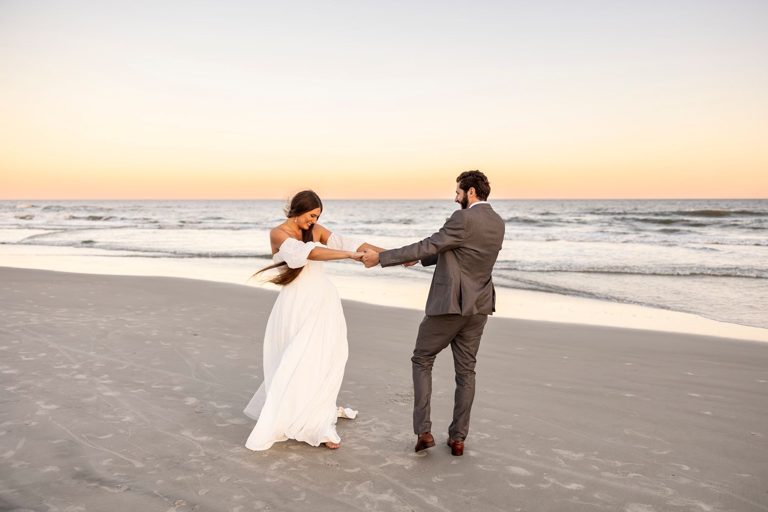A couple in wedding attire dances together on a sandy beach at sunset with ocean waves in the background.