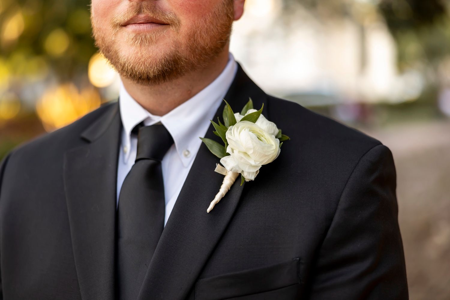 Close-up photo showing white rose boutonniere pinned to the lapel of a black formal suit jacket.