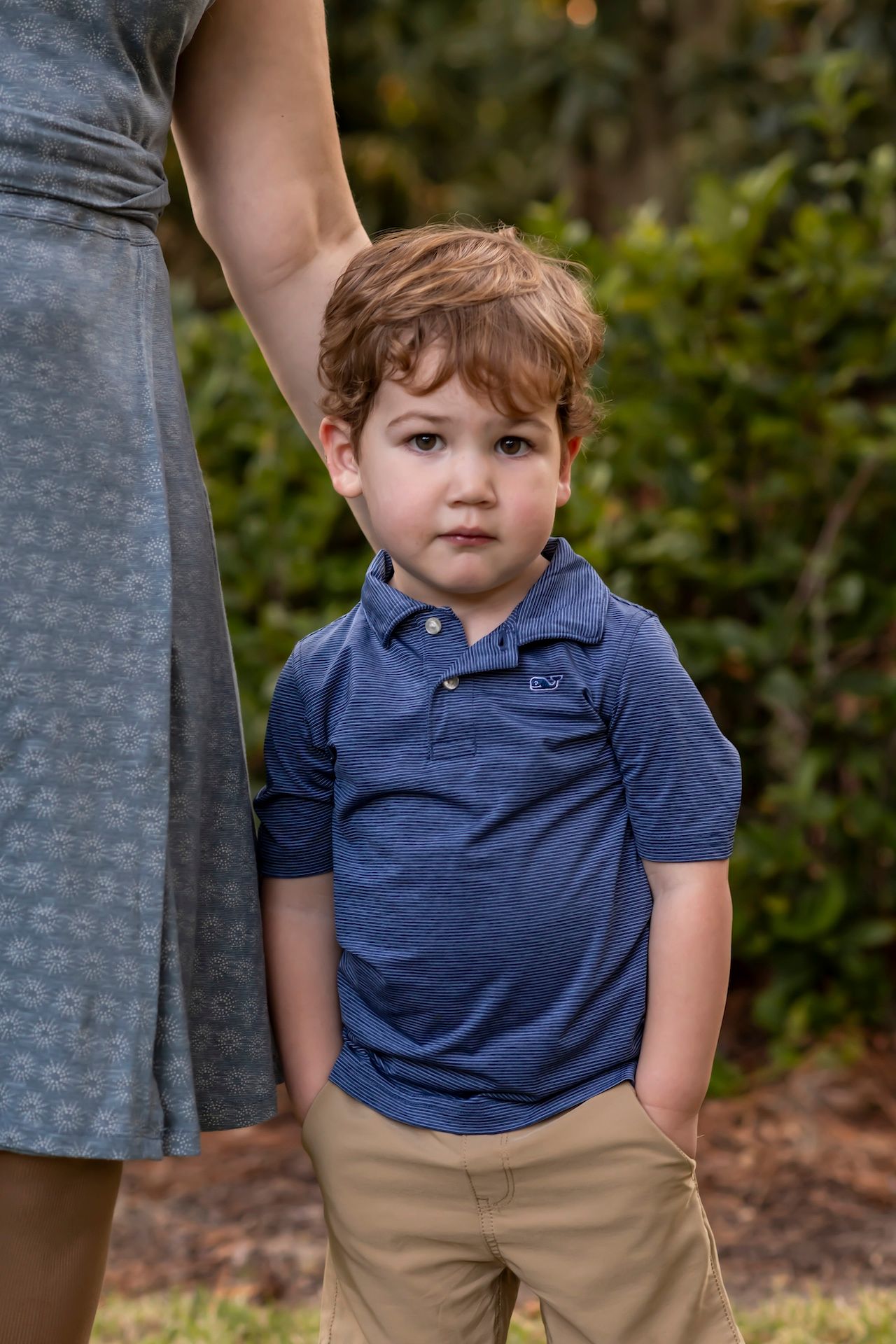 Small child in a navy polo shirt and khaki shorts standing next to adult wearing gray dress outdoors.
