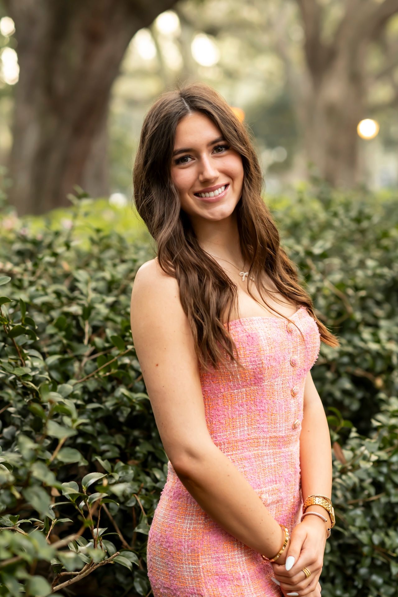 Someone in a coral pink patterned dress poses outdoors against green foliage with bokeh lights in the background.