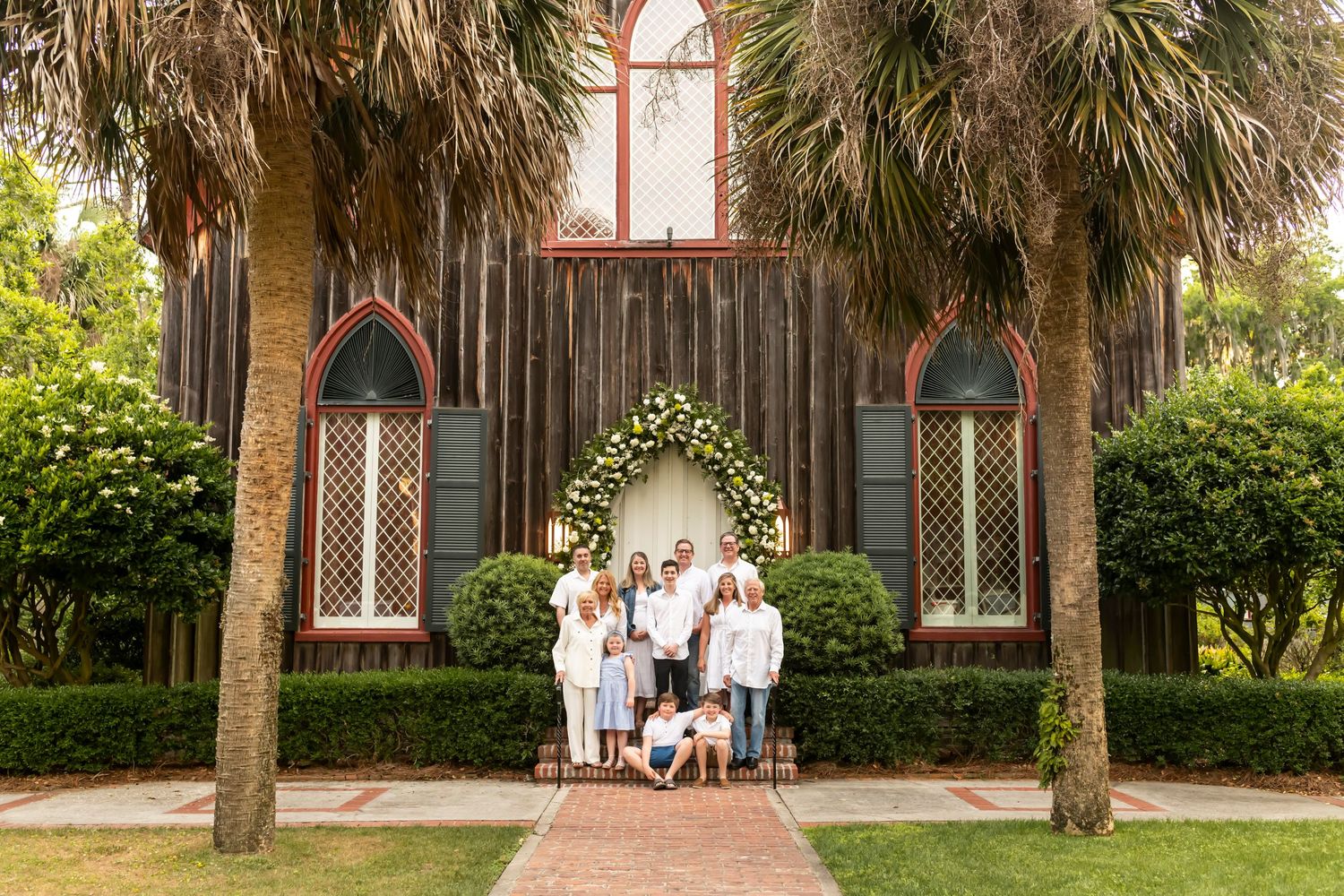 A group poses together under a floral arch in front of a rustic chapel with palm trees and gothic windows.