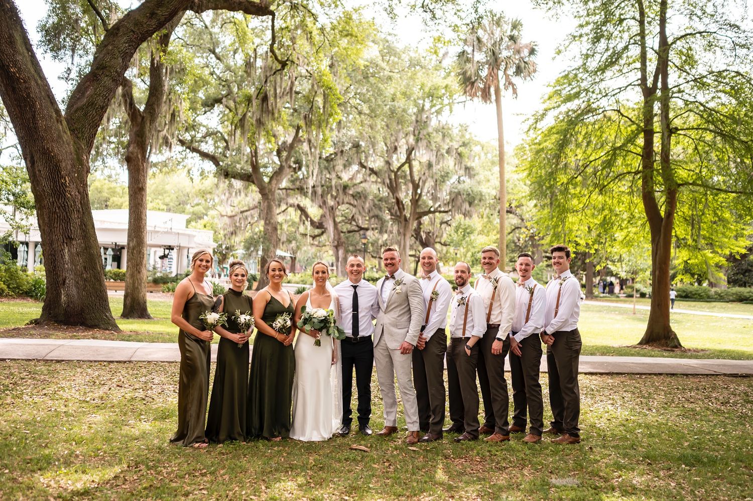 Wedding party in formal attire poses together under Spanish moss draped oak trees in a lush Southern garden setting.