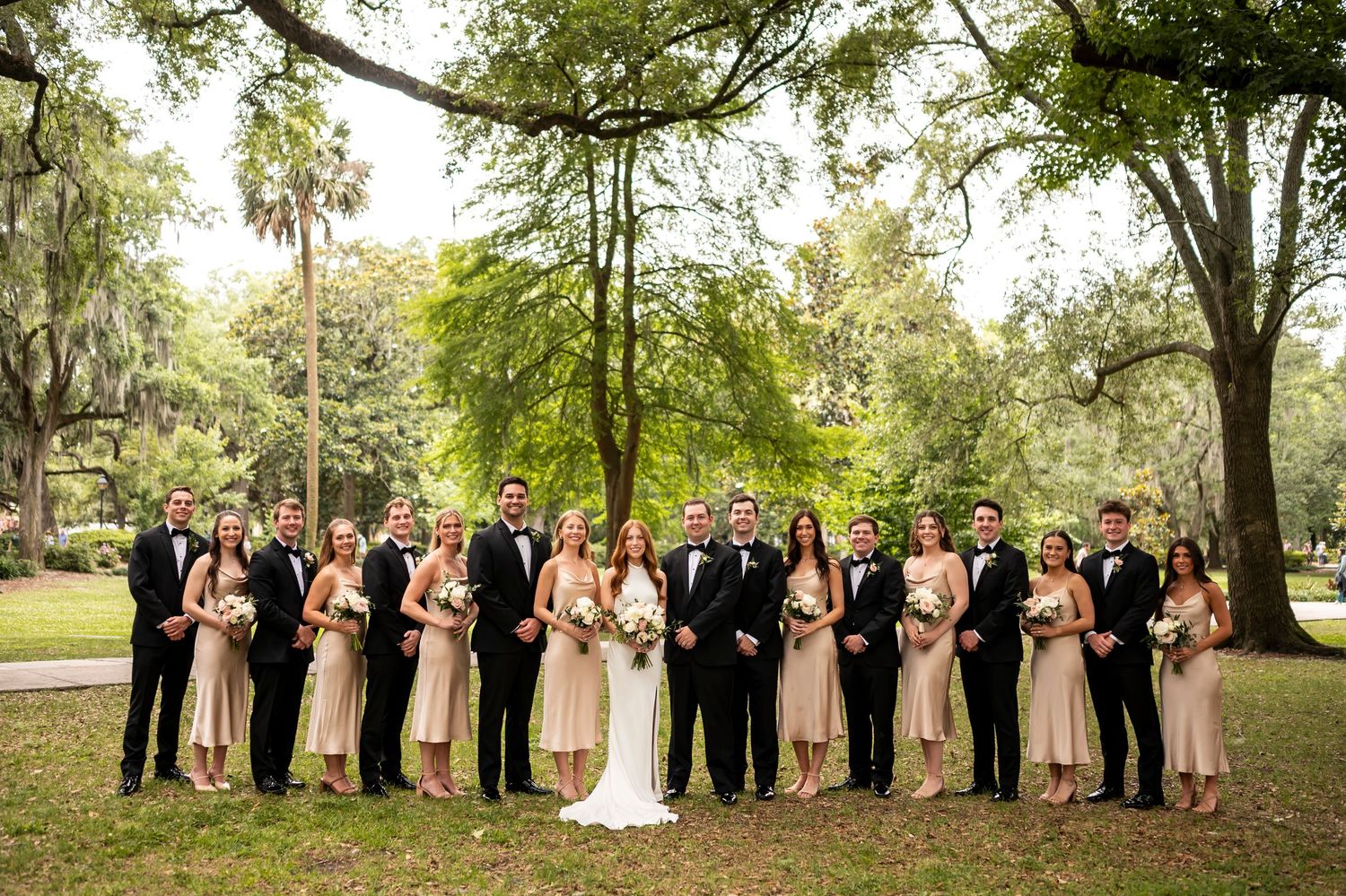 Wedding party in formal attire stands together under moss-draped trees in an elegant outdoor garden setting.
