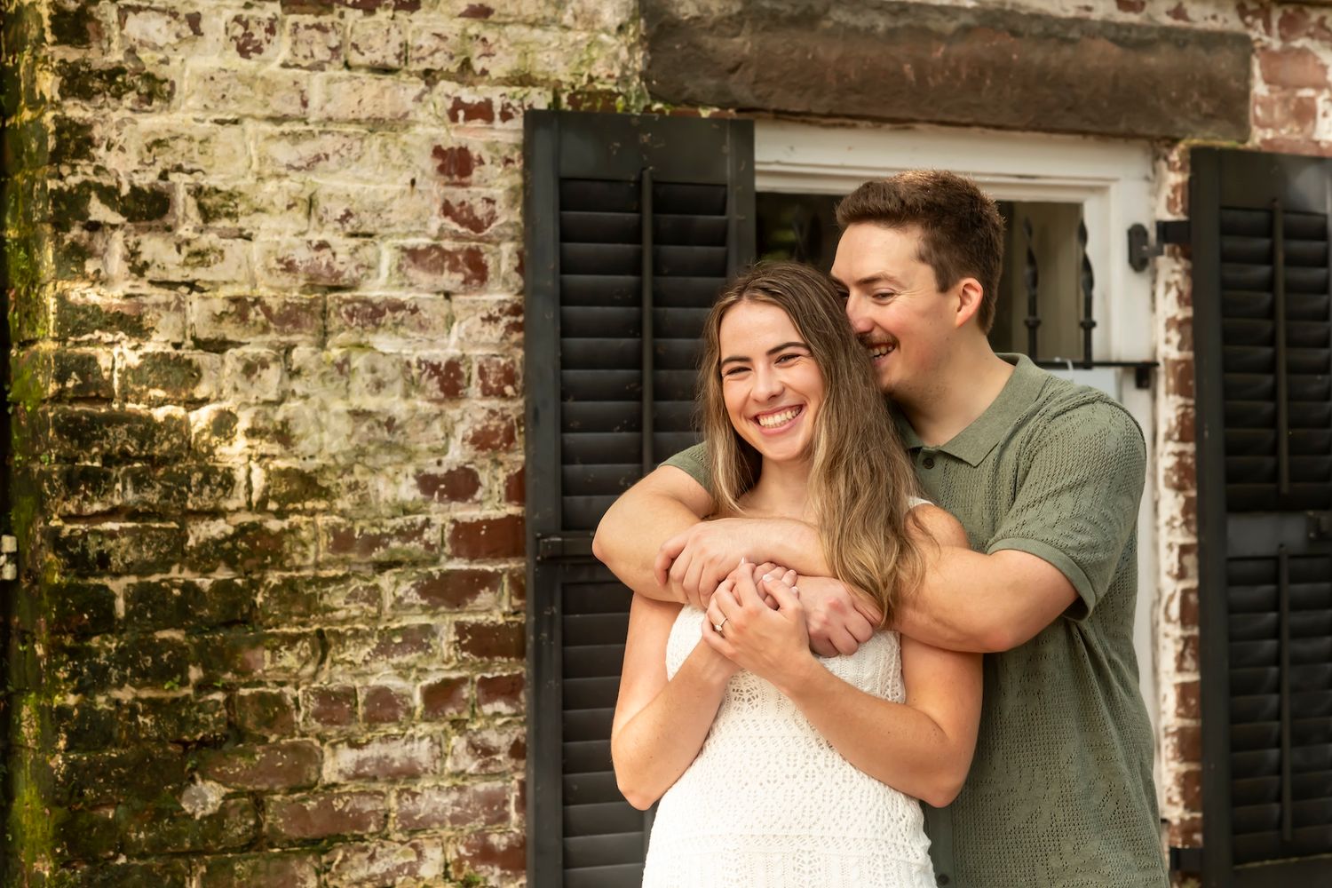 Two people embrace in a romantic pose against a rustic brick wall with black shutters in the background.